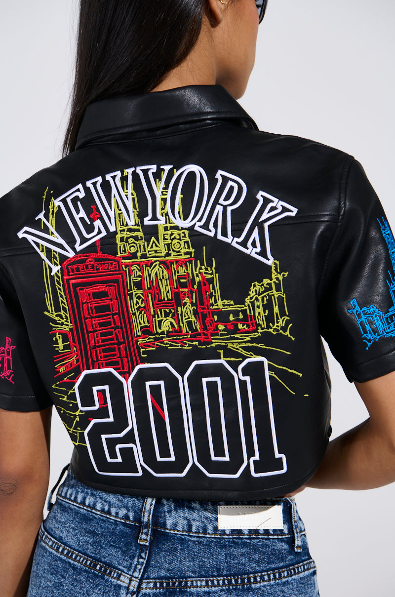 A person with long brown hair wearing blue jeans and the MAD CITY FAUX LEATHER SHORT SLEEVE BUTTON DOWN SHIRT, featuring large "NEW YORK" and "2001" embroidery. The back displays colorful line art of buildings, including a red phone booth, creating a cropped silhouette.