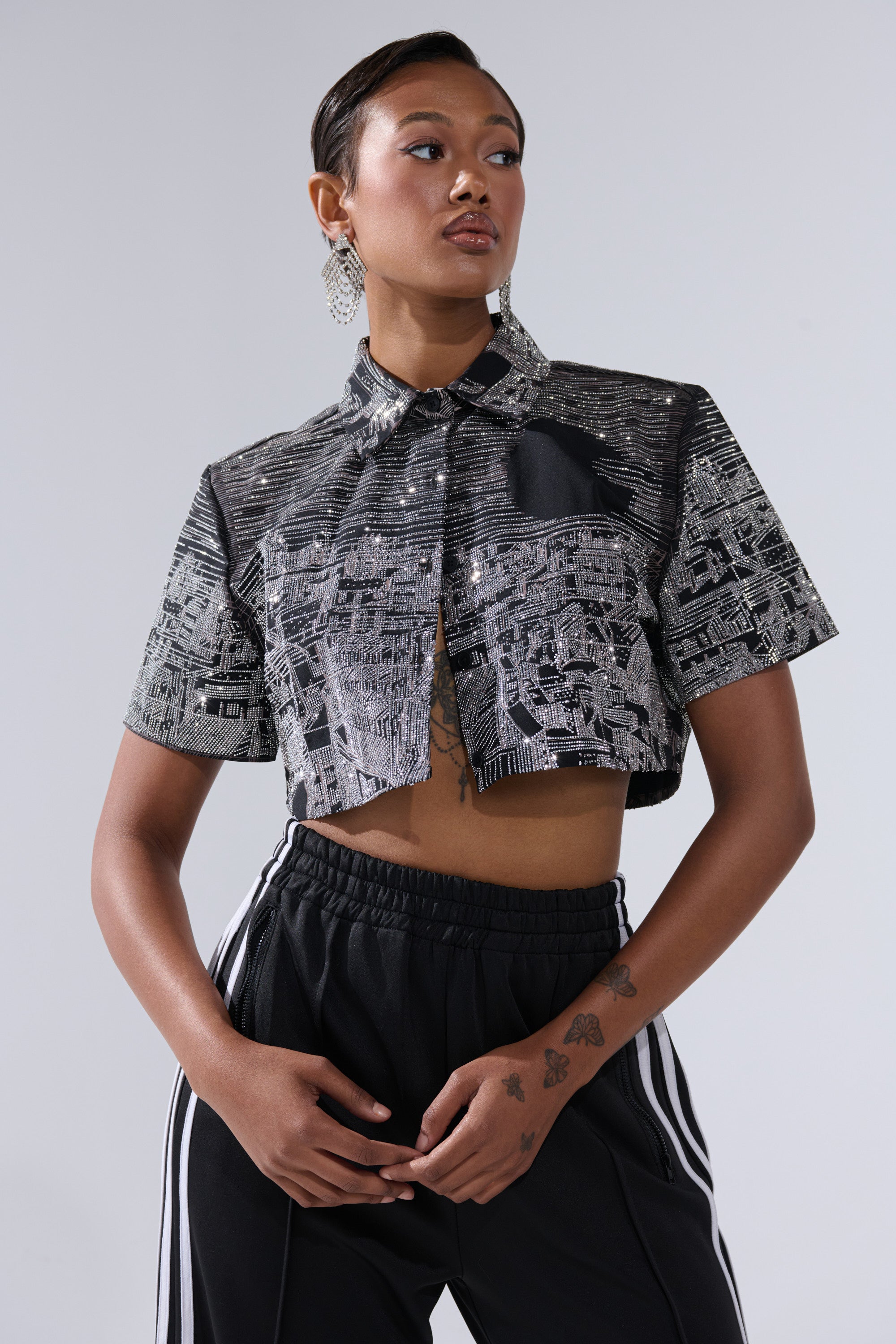 A woman poses confidently in the ALL AROUND THE WORLD BUTTON DOWN TOP IN BLACK, styled with black track pants with white stripes. She accessorizes with bold earrings and displays tattoos on her left hand.