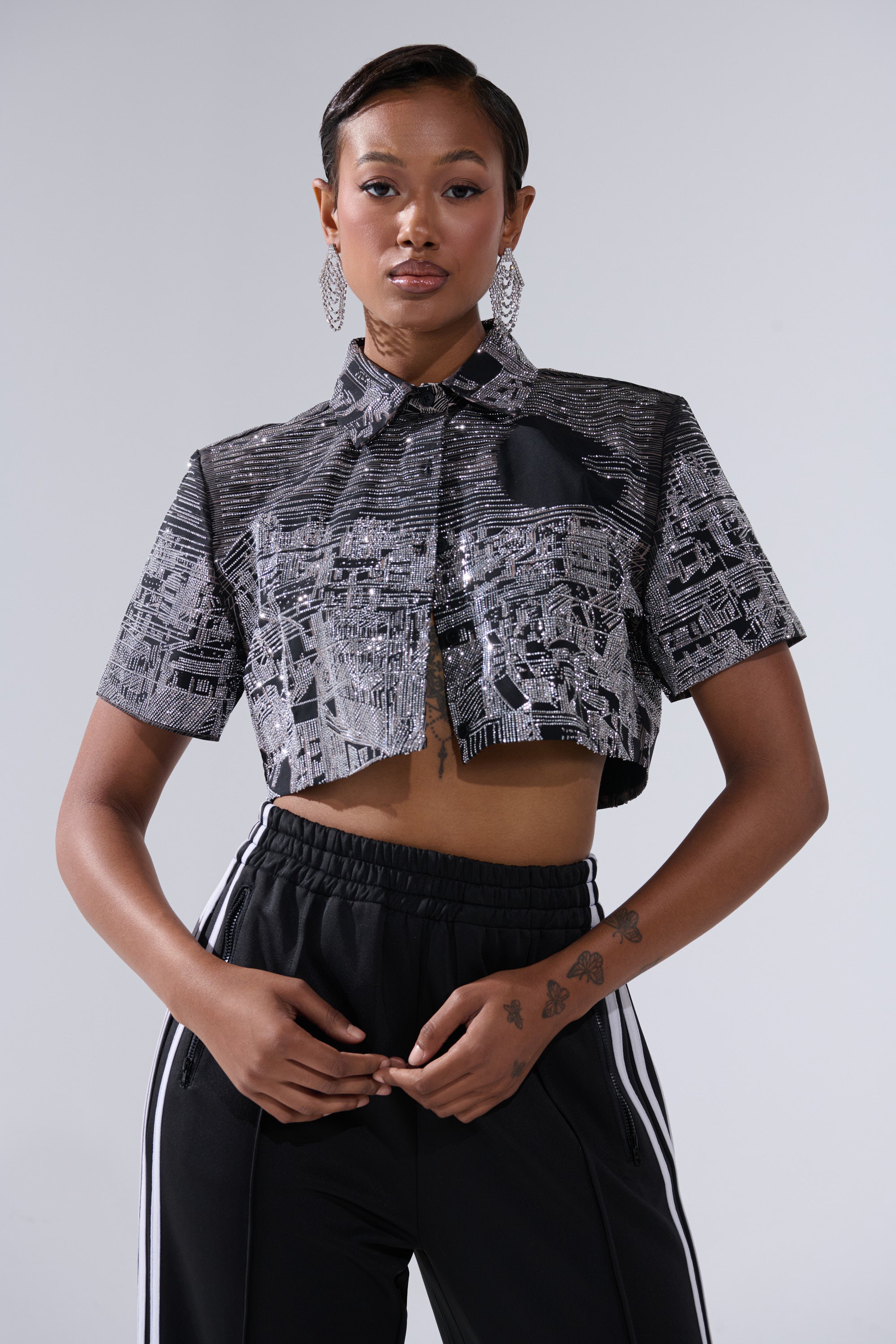 A woman poses confidently in the ALL AROUND THE WORLD BUTTON DOWN TOP IN BLACK, styled with black track pants with white stripes and large dangling earrings, against a plain light gray background.