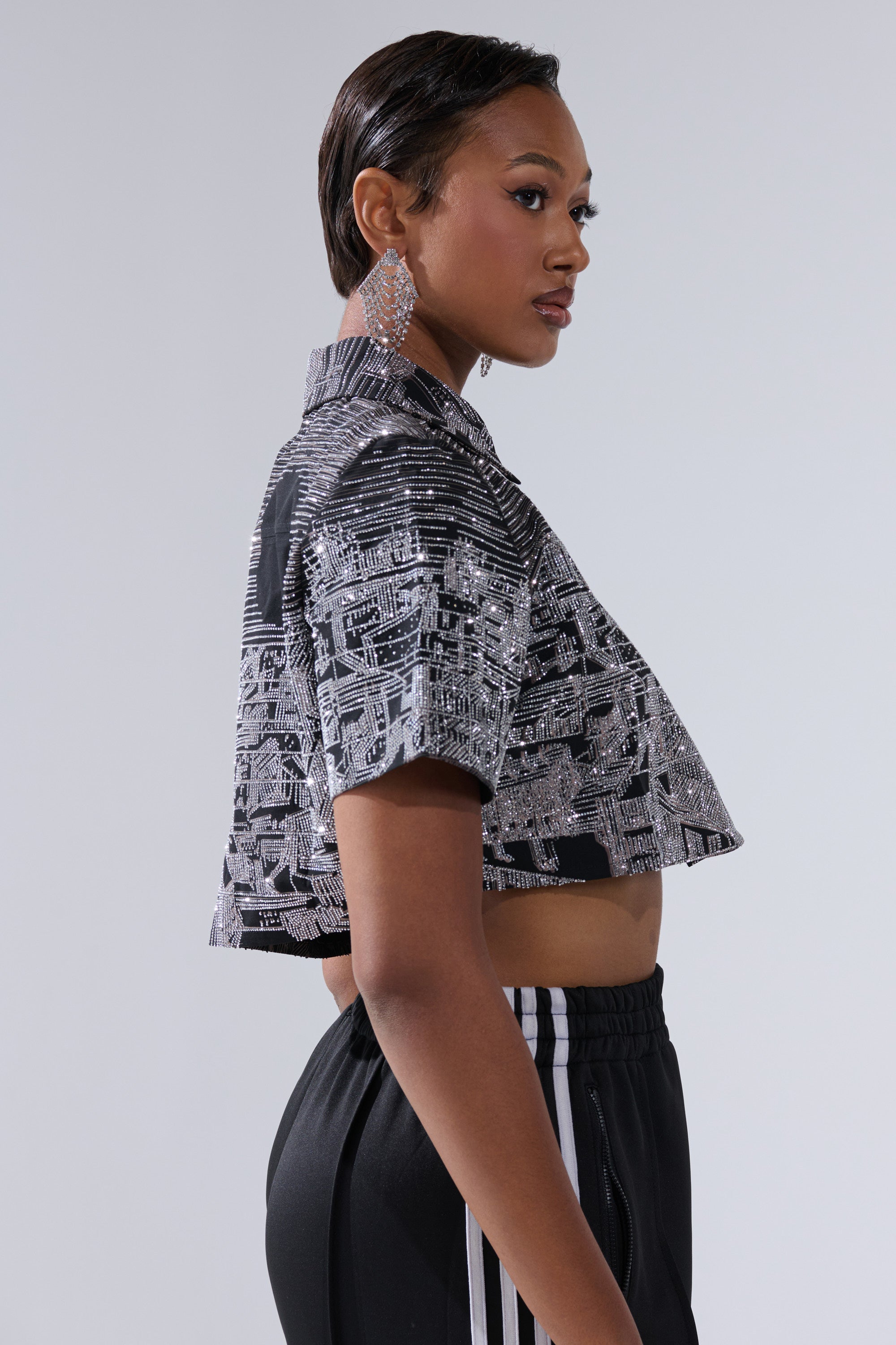 A woman with short hair, wearing the ALL AROUND THE WORLD BUTTON DOWN TOP IN BLACK, black pants with white side stripes, and large statement earrings, poses in profile against a plain light background.