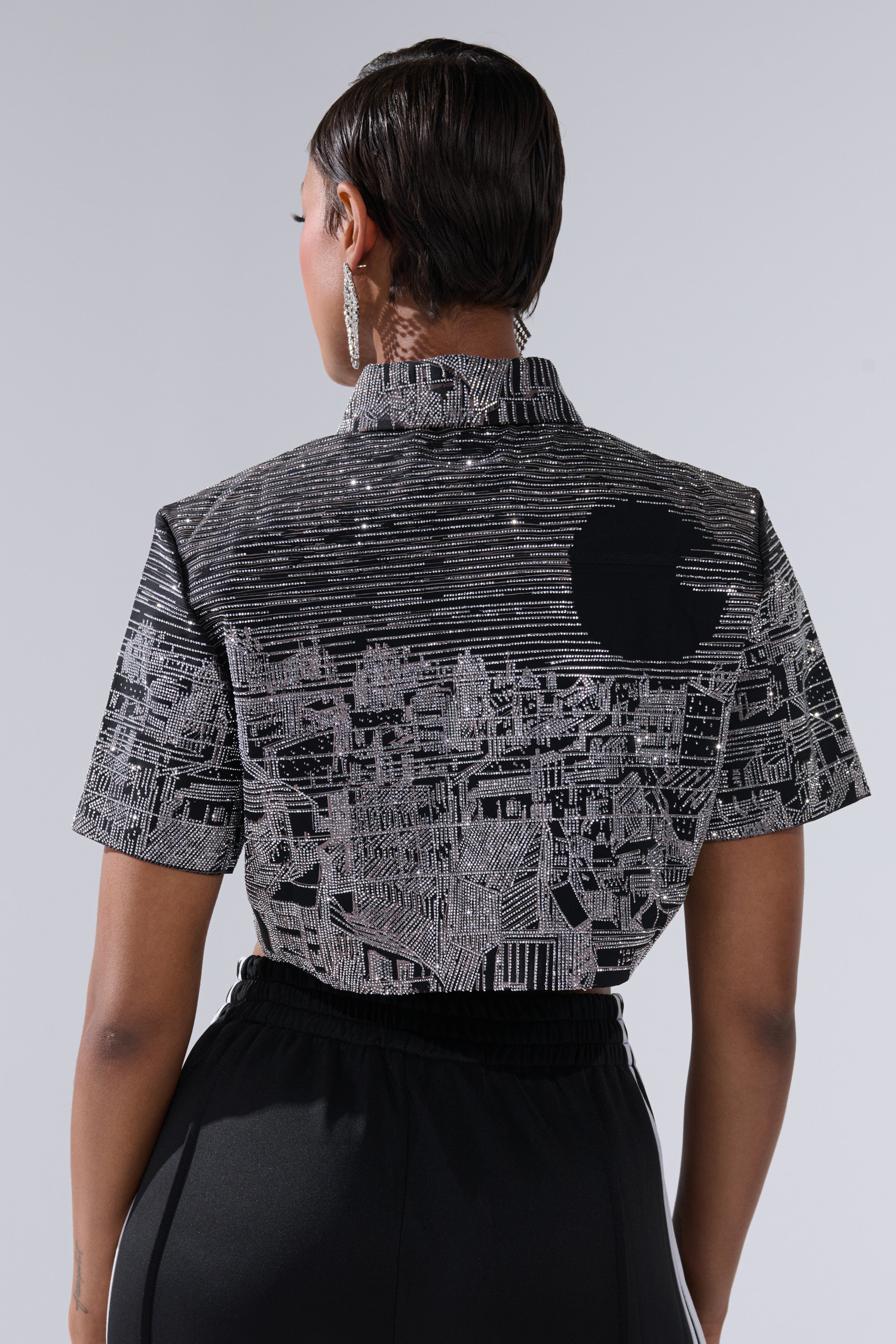 A person with short hair, seen from behind, wears the ALL AROUND THE WORLD BUTTON DOWN TOP IN BLACK—featuring a cityscape graphic and large black circle—paired with black pants against a plain light gray background.