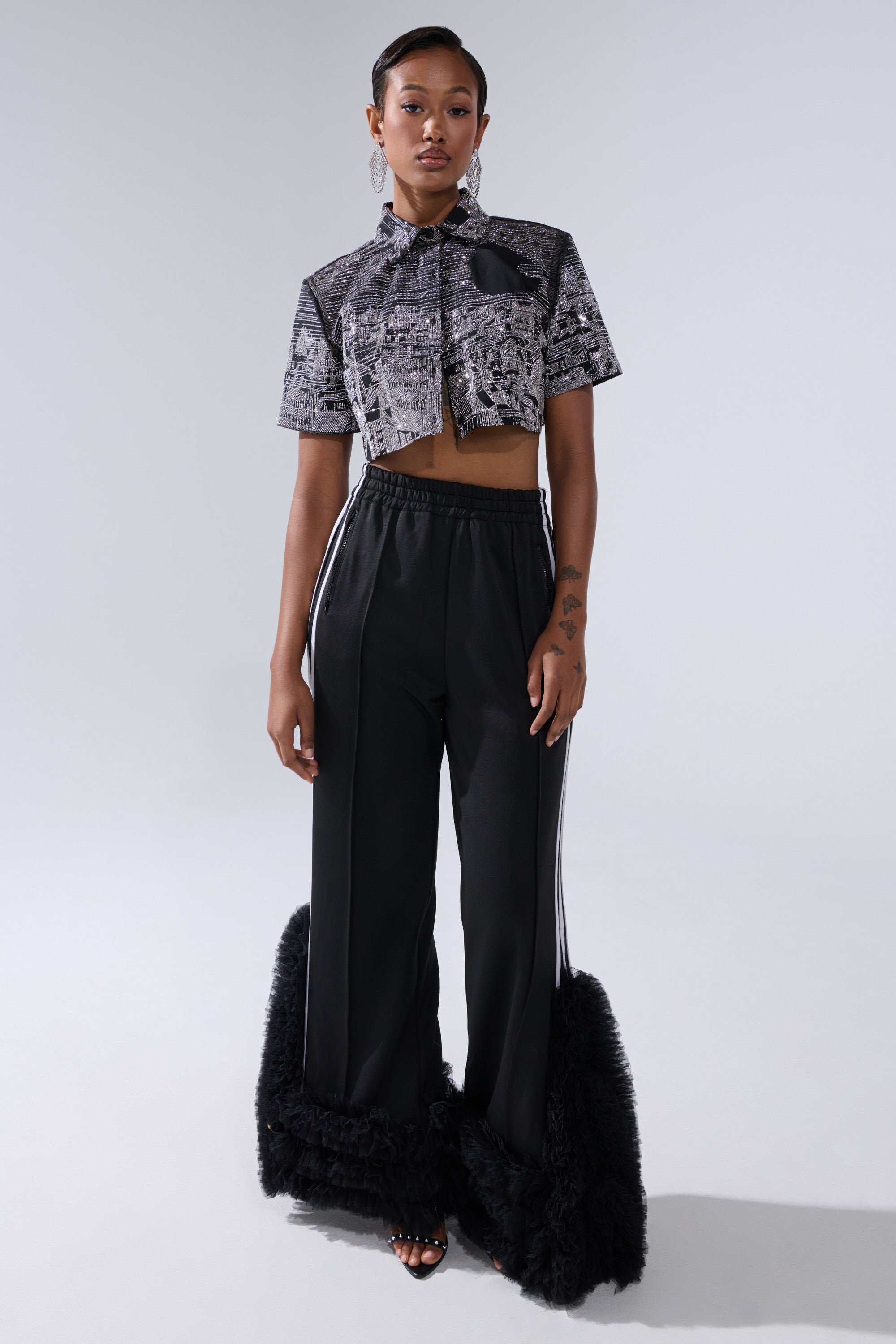 A woman with short hair and a confident expression wears the ALL AROUND THE WORLD BUTTON DOWN TOP IN BLACK, styled with high-waisted black pants featuring wide, fringed hems and open-toed heels, standing against a plain background.