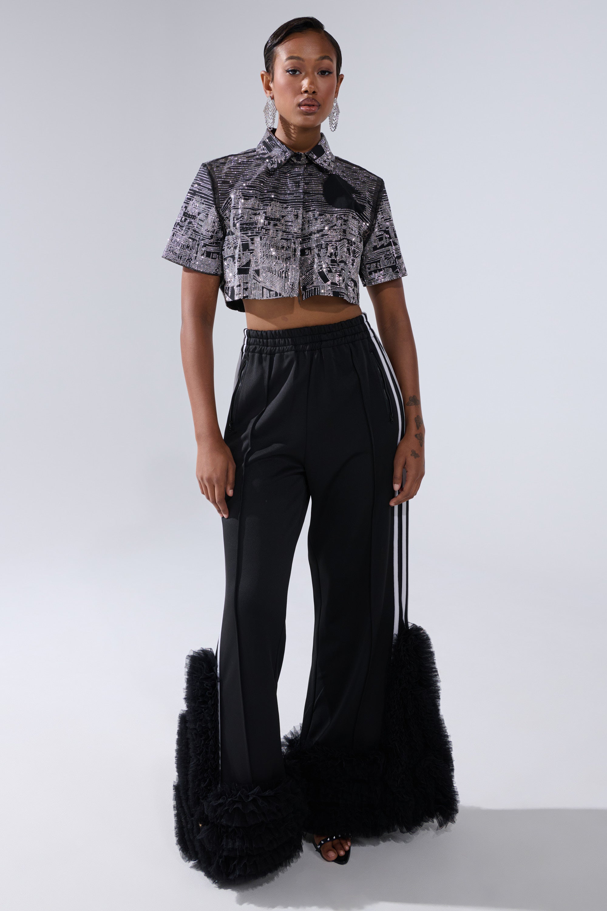 Against a light gray background, a woman wears the ALL AROUND THE WORLD BUTTON DOWN TOP IN BLACK, paired with high-waisted black pants with wide, ruffled hems, open-toe black heels, and drop earrings.