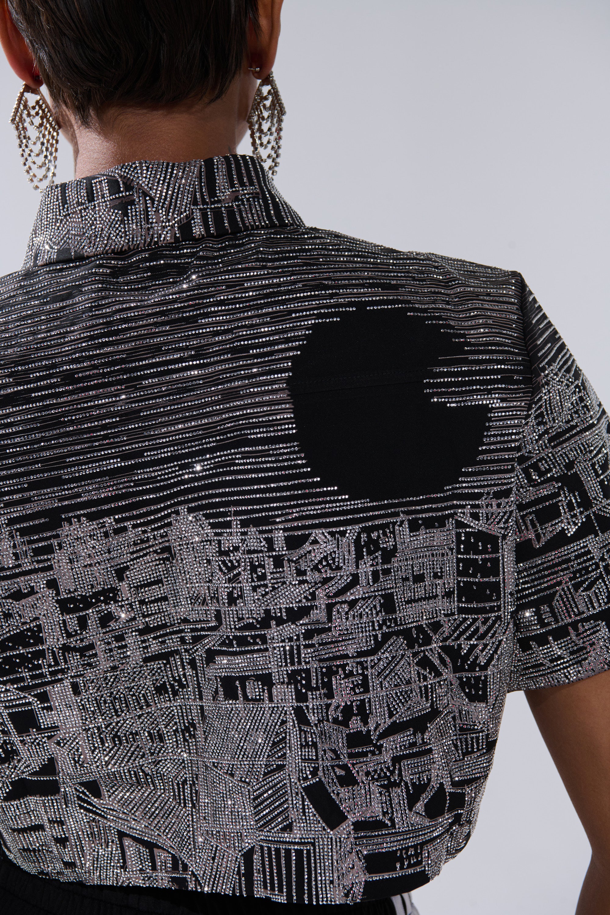 Close-up of a person wearing the ALL AROUND THE WORLD BUTTON DOWN TOP IN BLACK, featuring cropped short sleeves, a shimmering silver cityscape graphic, and a bold black circle on the back. They have short hair and sparkly earrings.