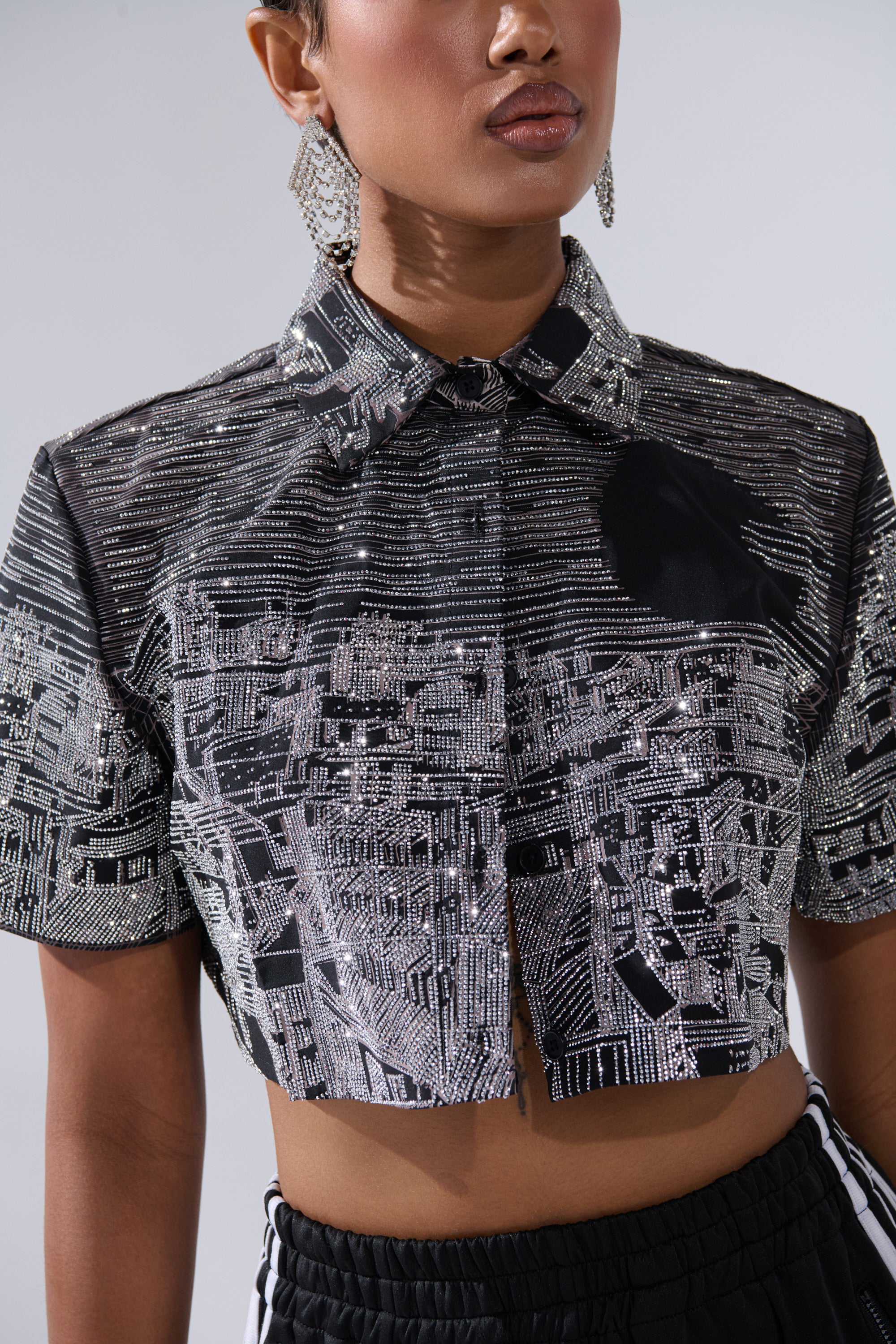 A person wears the ALL AROUND THE WORLD BUTTON DOWN TOP IN BLACK—a cropped, sequin-embellished design—paired with dangling earrings and black pants with white stripes. Their face is partially out of frame.