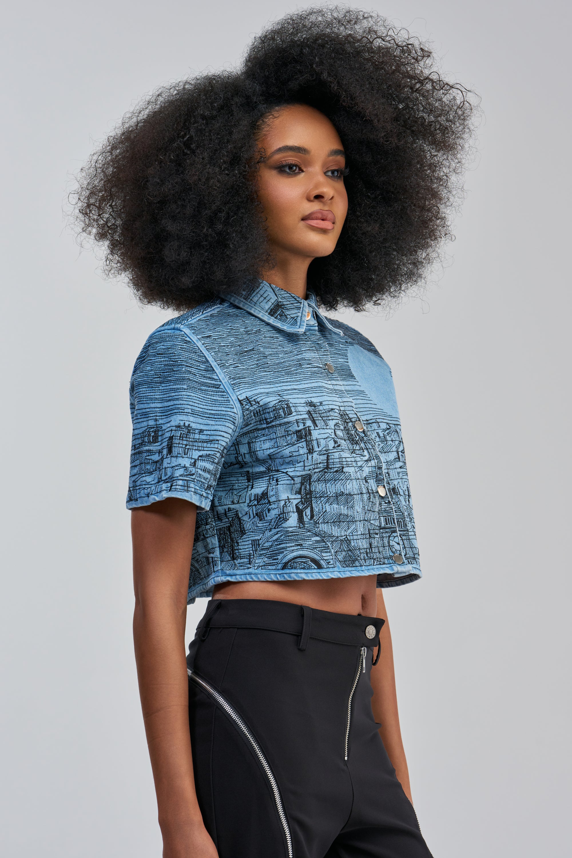 A model with a voluminous afro hairstyle wears the INK HORIZON EMBROIDERED TOP IN LIGHT BLUE DENIM, a short-sleeved, cropped button-down shirt embellished with a black sketched cityscape pattern, paired with black streetwear-inspired pants featuring silver zippers along the thighs, set against a plain gray background.