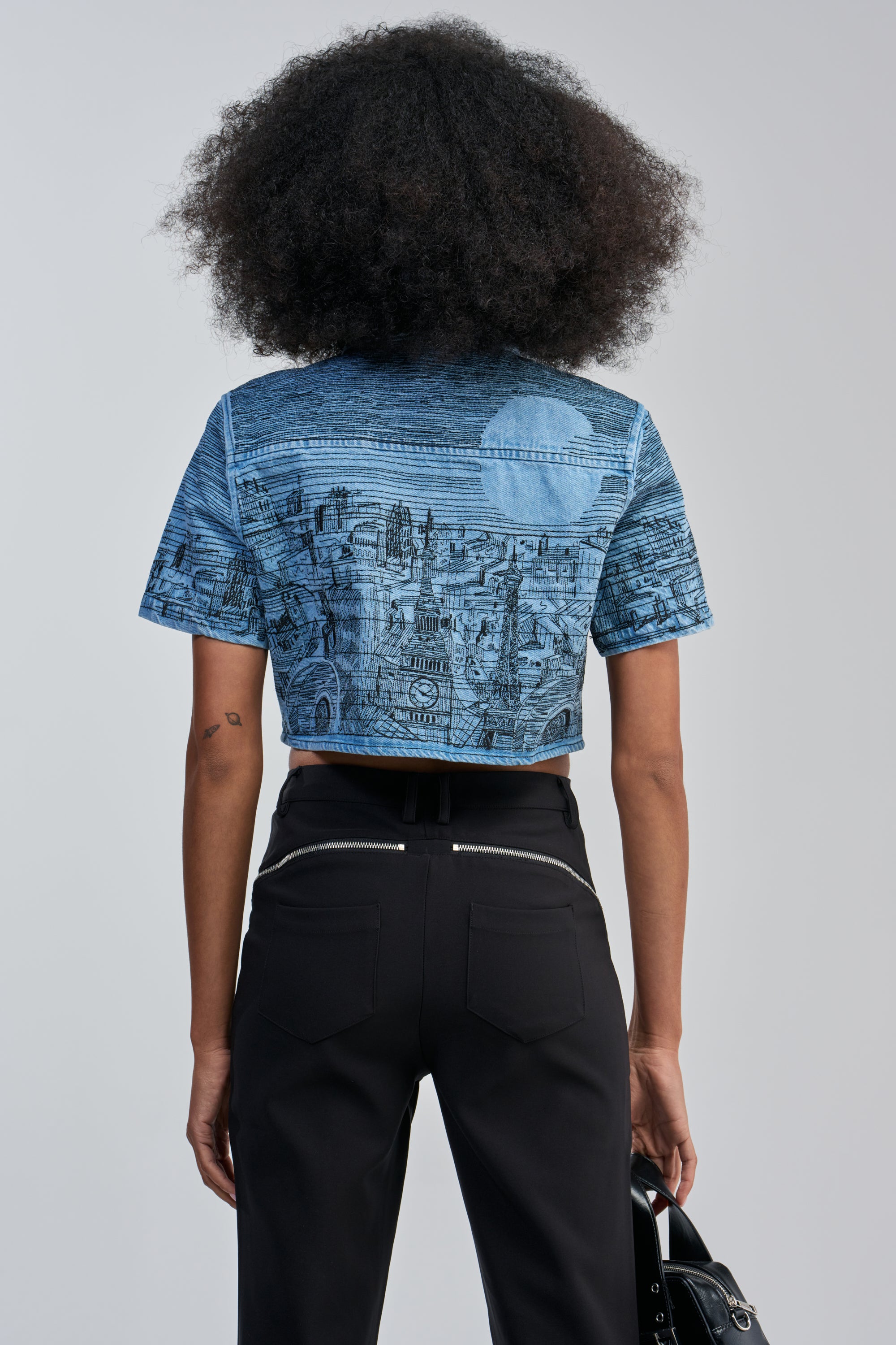 A person with curly hair, seen from behind, is wearing the INK HORIZON EMBROIDERED TOP IN LIGHT BLUE DENIM styled as streetwear with black zipper-accented pants and holding a black bag.