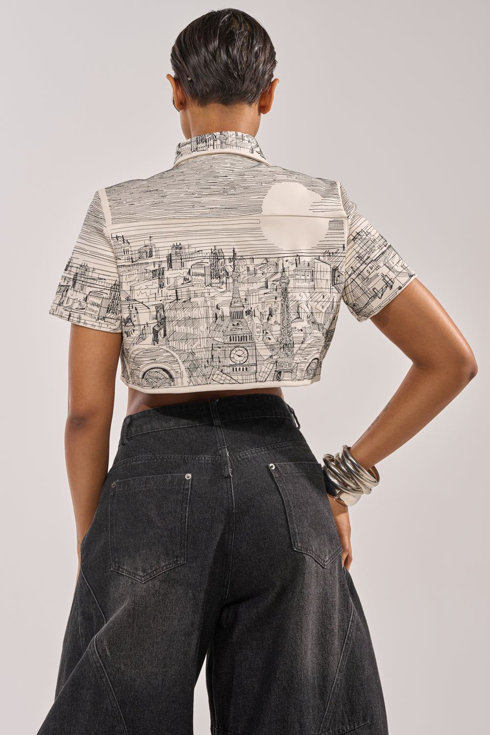 A person with short dark hair stands with their back to the camera, wearing the INK HORIZON EMBROIDERED TOP—a faux leather cropped piece adorned with cityscape embroidery—and high-waisted black jeans, against a plain background. 5
