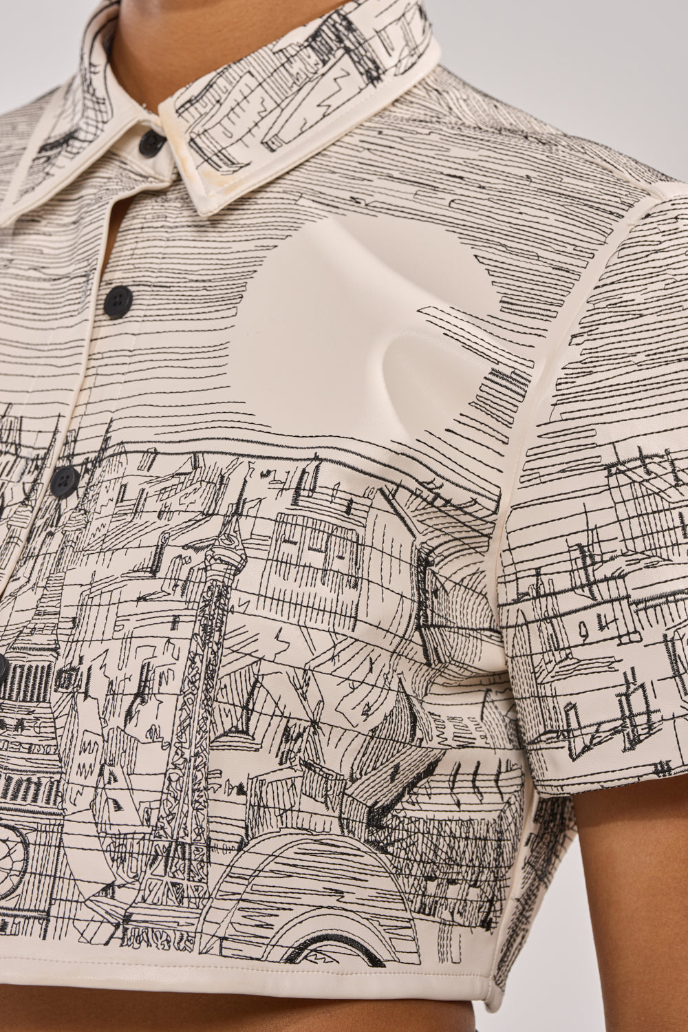 A close-up of someone wearing the INK HORIZON EMBROIDERED TOP, a beige shirt adorned with black cityscape embroidery of landmarks, architectural details, and a large sun or moon in the background. 7