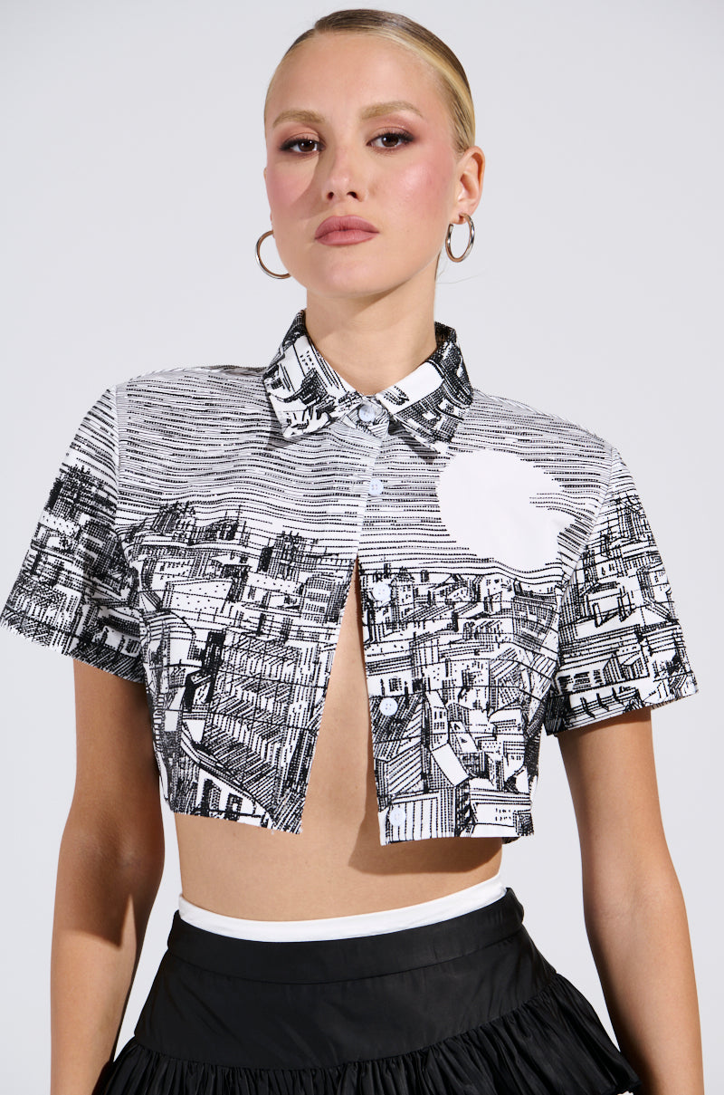 A person with short blonde hair is wearing the ALL AROUND THE WORLD BUTTON DOWN TOP in black and white, featuring a cityscape graphic, paired with a ruffled black skirt against a plain gray background that enhances the outfit's details. 1