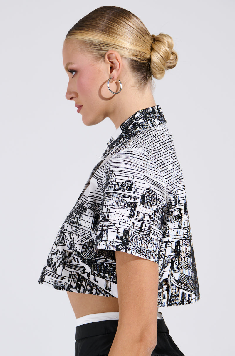 A person with a sleek bun wears the AKIRA Label "ALL AROUND THE WORLD BUTTON DOWN TOP," a black and white cropped shirt showcasing an intricate cityscape graphic. Hoop earrings dangle as they're photographed in side profile against a plain background. 2