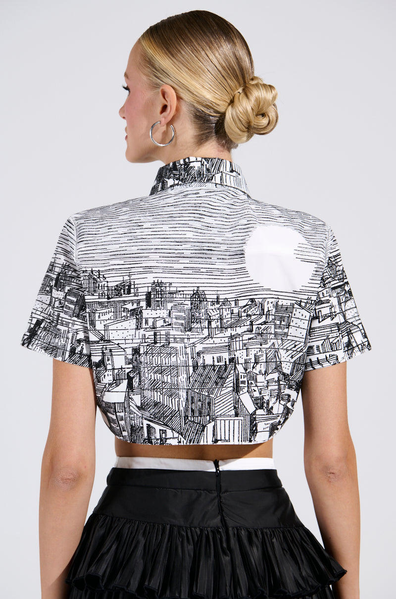 A woman with blonde hair in a bun wears the "All Around The World Button Down Top" with a cityscape graphic, paired with a black tiered skirt. She is slightly turned, showcasing the intricate design of her outfit. 3