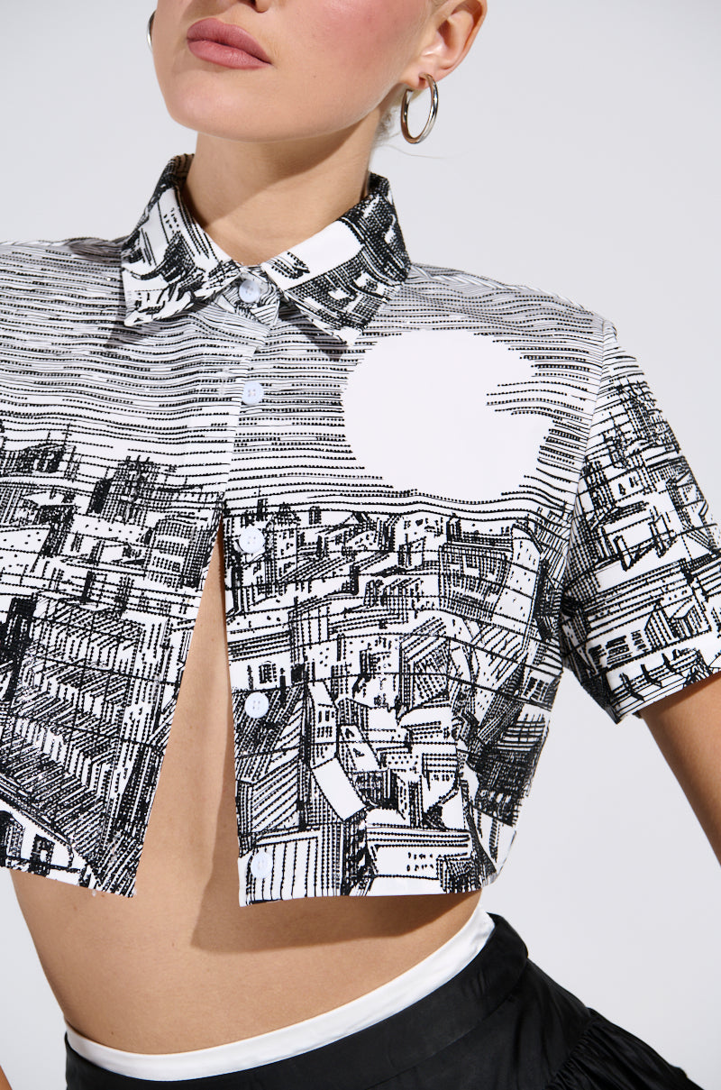 Sporting the "ALL AROUND THE WORLD BUTTON DOWN TOP" with its black and white cityscape graphic and chic cut-out design, the individual exudes effortless style. Their hoop earrings enhance the urban chic vibe against a plain backdrop. 6