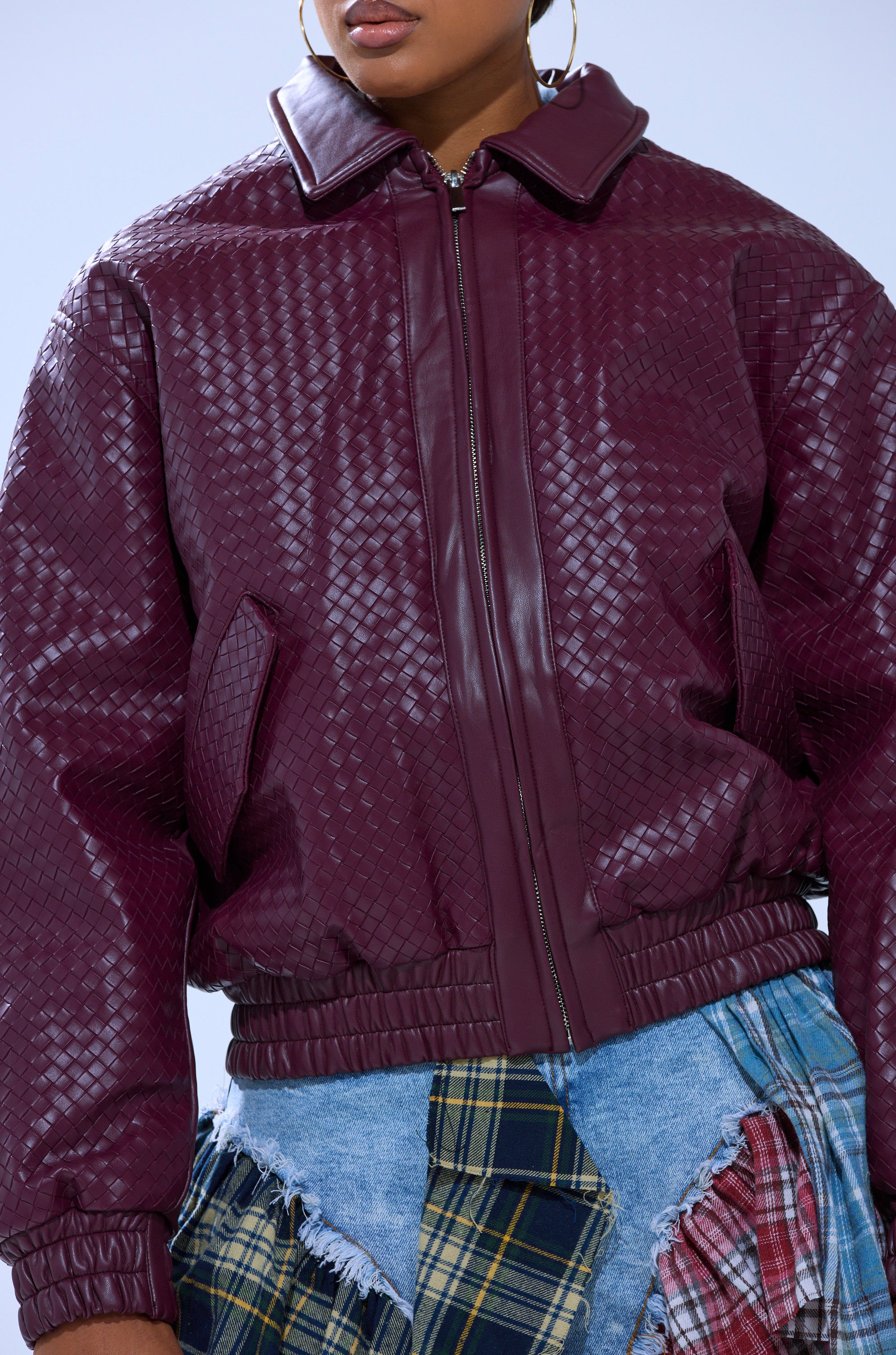 A person wears the CHERRY POP BOMBER, an oversized maroon textured zip-up jacket with a collar and elastic cuffs, paired with a patchwork denim skirt in various plaid patterns. The person’s face is partially out of frame.