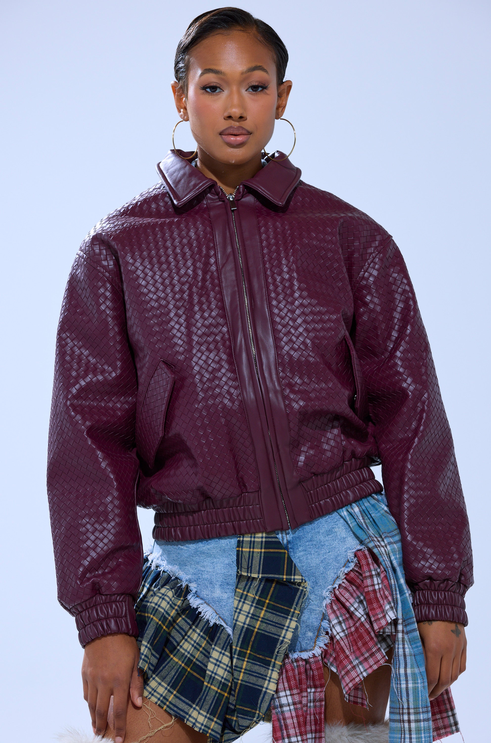 Against a light blue background, a woman wears the CHERRY POP BOMBER faux leather jacket with a patchwork denim skirt featuring plaid accents. She accessorizes with large hoop earrings and has her hair slicked back.