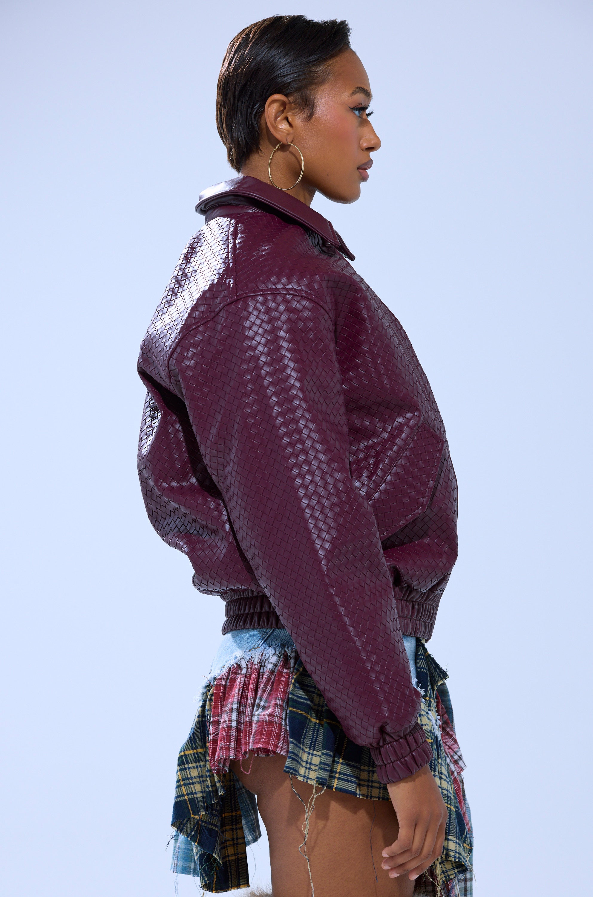 A person in profile models the CHERRY POP BOMBER with large hoop earrings and a short, multi-colored plaid skirt with frayed edges against a light blue background.