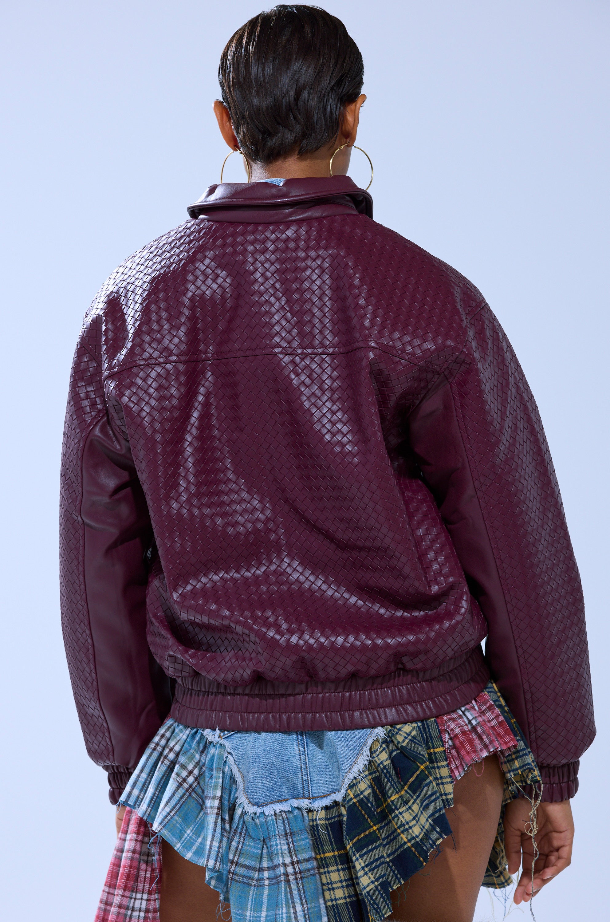 Seen from behind, a person with short dark hair wears the CHERRY POP BOMBER, featuring a shiny textured finish, paired with a blue plaid skirt with denim details and frayed edges against a light background.