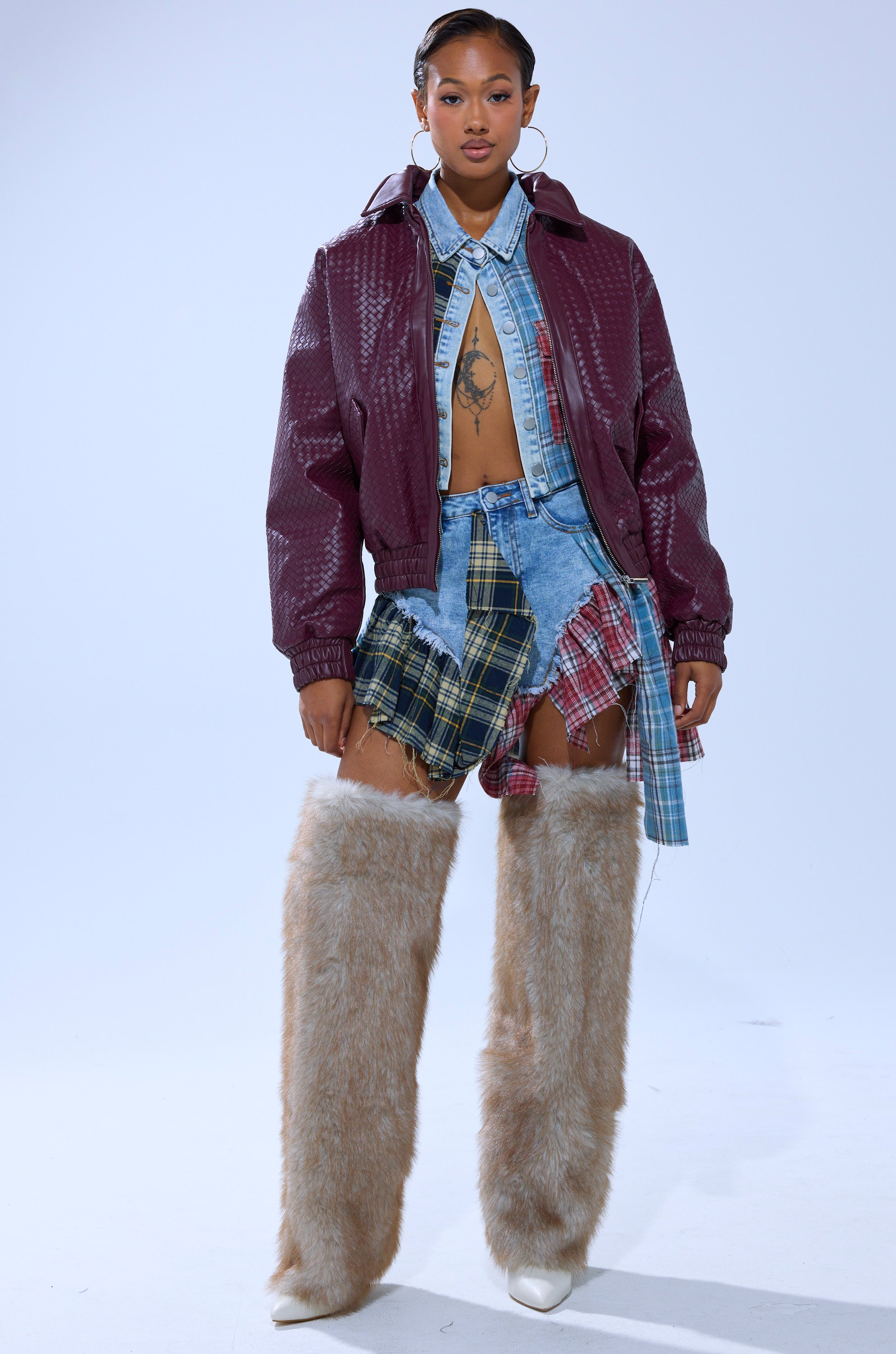 A woman poses confidently in the CHERRY POP BOMBER over a denim shirt and skirt with plaid patches, paired with tall furry beige boots. She accessorizes with hoop earrings and wears her hair styled back.