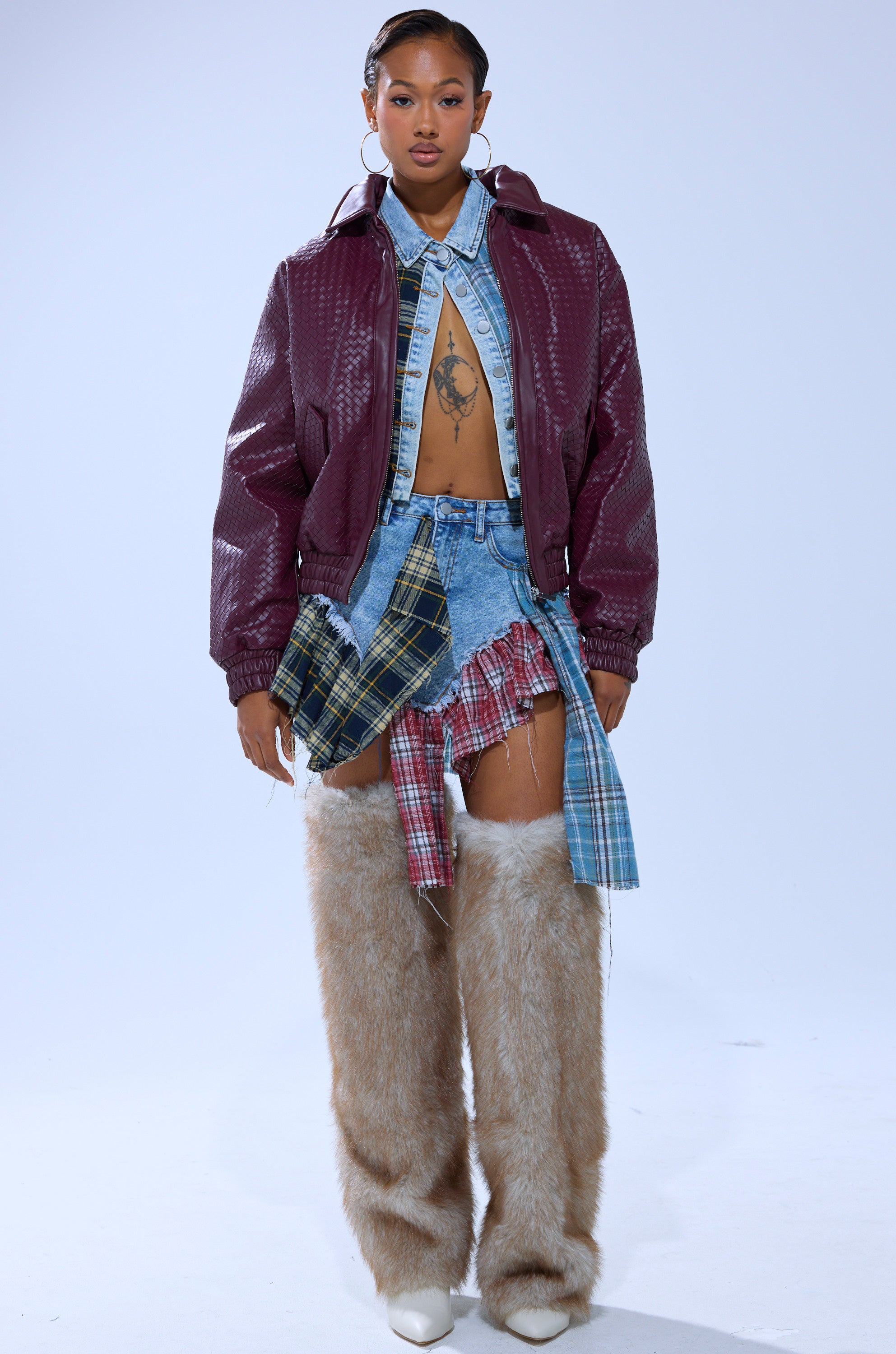 A person stands facing forward, wearing the CHERRY POP BOMBER, an unbuttoned denim shirt, a patchwork plaid skirt layered over denim shorts, tall furry beige leg warmers, white shoes, and their hair pulled back.