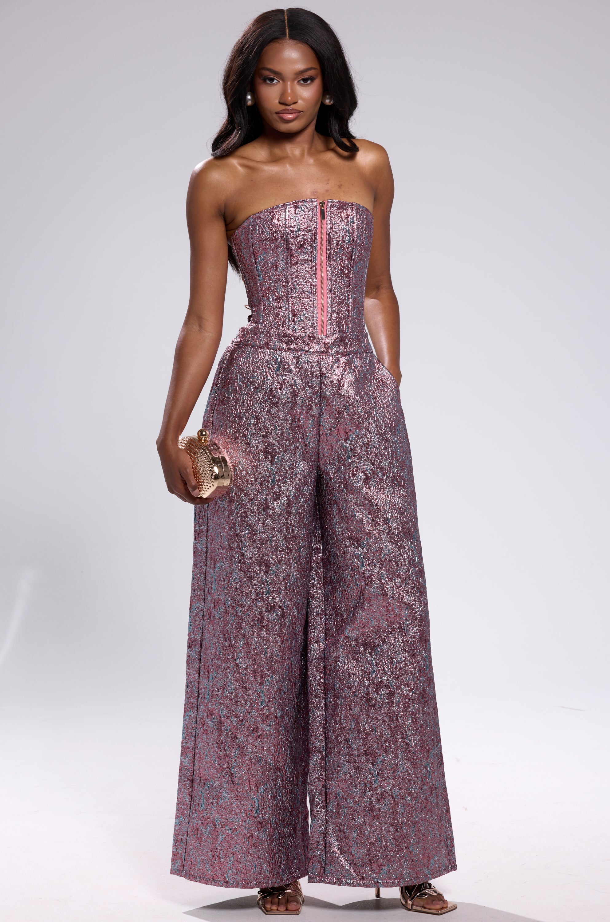 A woman stands confidently in NO COMPETITION BROCADE TROUSER—strapless, metallic pink with high-rise wide-leg pants—holding a gold clutch. She has long, wavy hair and poses against a plain light background.