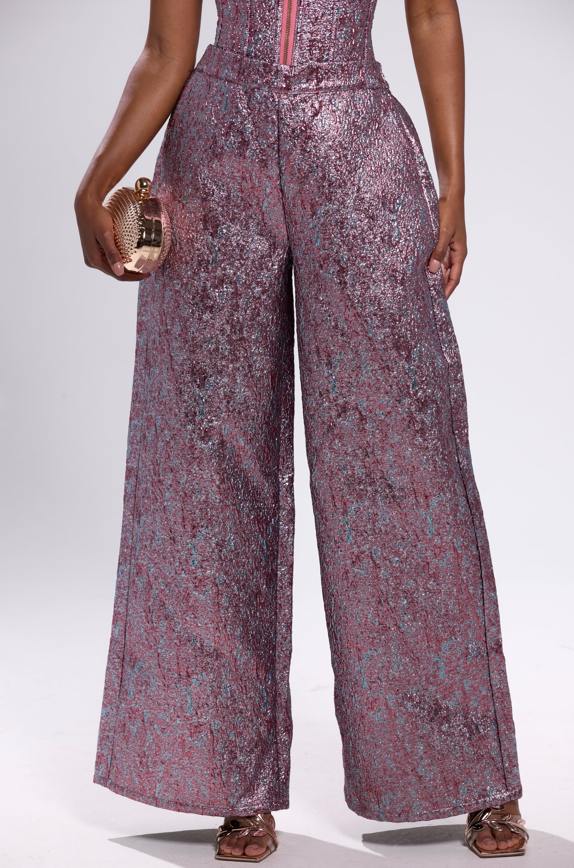 Dressed in NO COMPETITION BROCADE TROUSER—shiny pink metallic, wide-leg pants—a person stands against a white background, holding a round metallic clutch and wearing open-toed sandals.