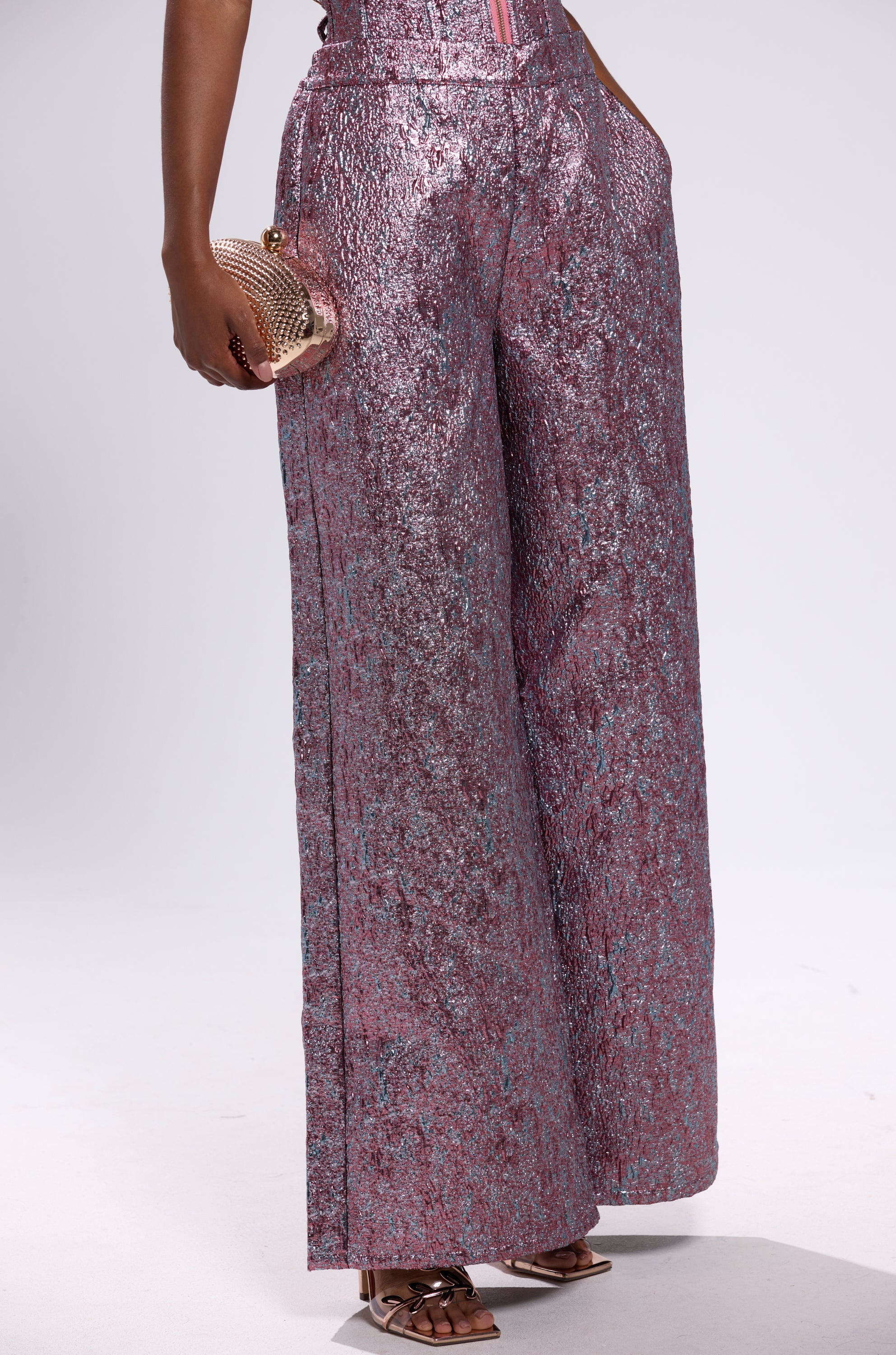 A person wears the NO COMPETITION BROCADE TROUSER—high-waisted, wide-leg metallic pink and purple pants with a textured finish—styled with a small gold mesh clutch and metallic open-toe heels against a plain white background.