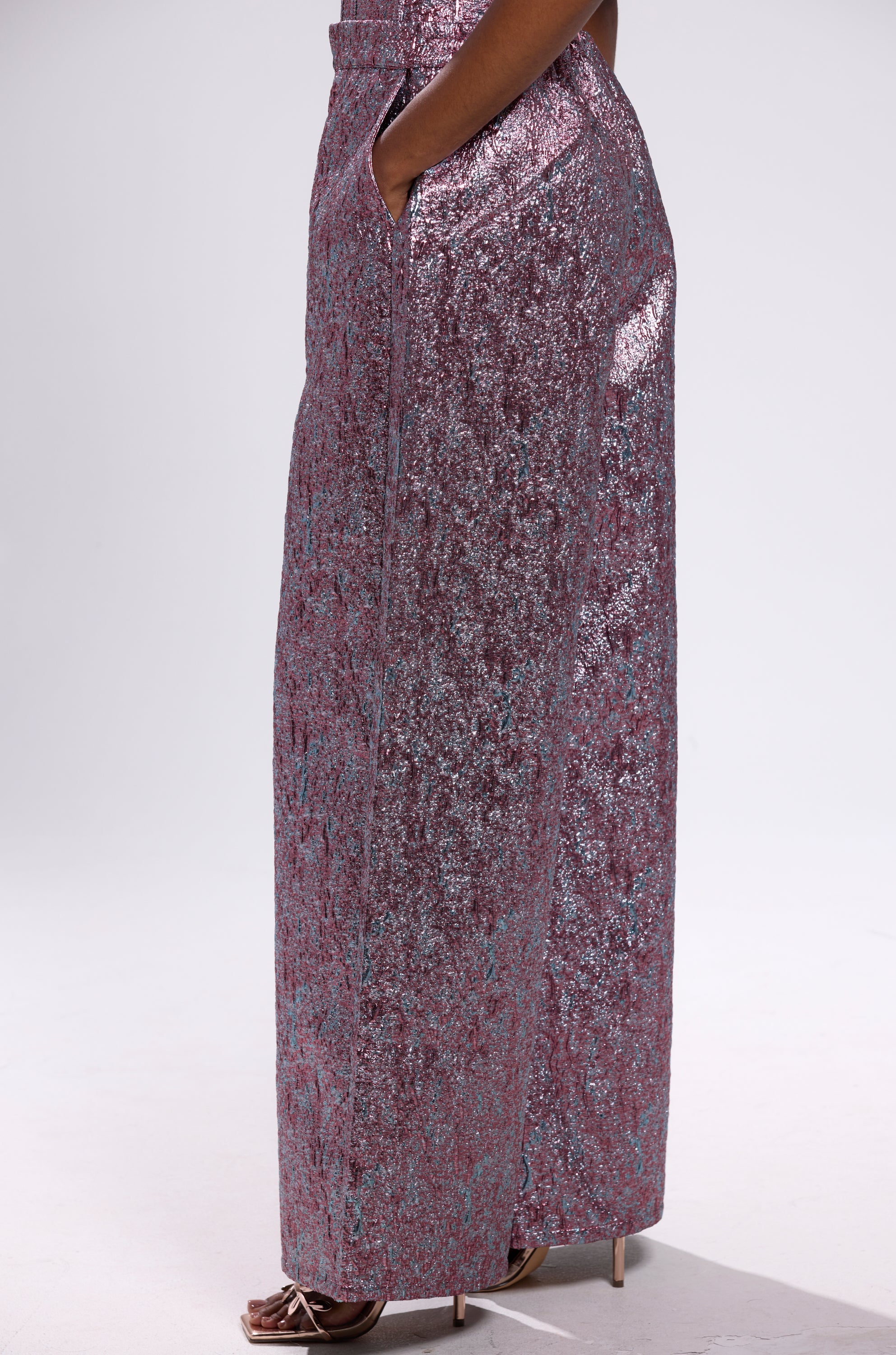A person stands sideways wearing NO COMPETITION BROCADE TROUSER—high-waisted, wide-leg trousers in shimmery pink and purple. With hands in pockets and strappy high heels, they complete a chic, standout look.