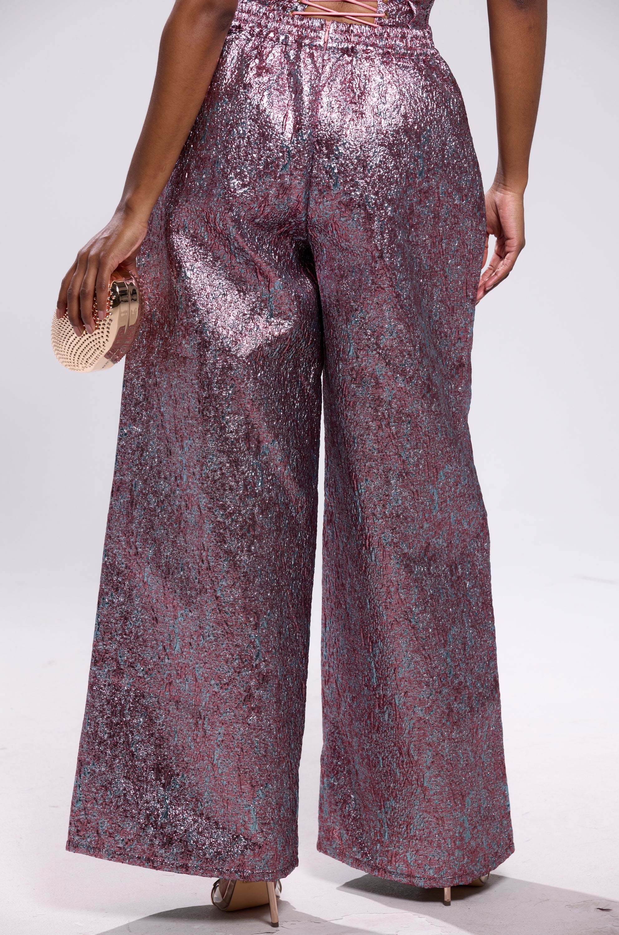 A person faces away, wearing the NO COMPETITION BROCADE TROUSER in metallic pink and silver, paired with high heels and holding a small, round light clutch.
