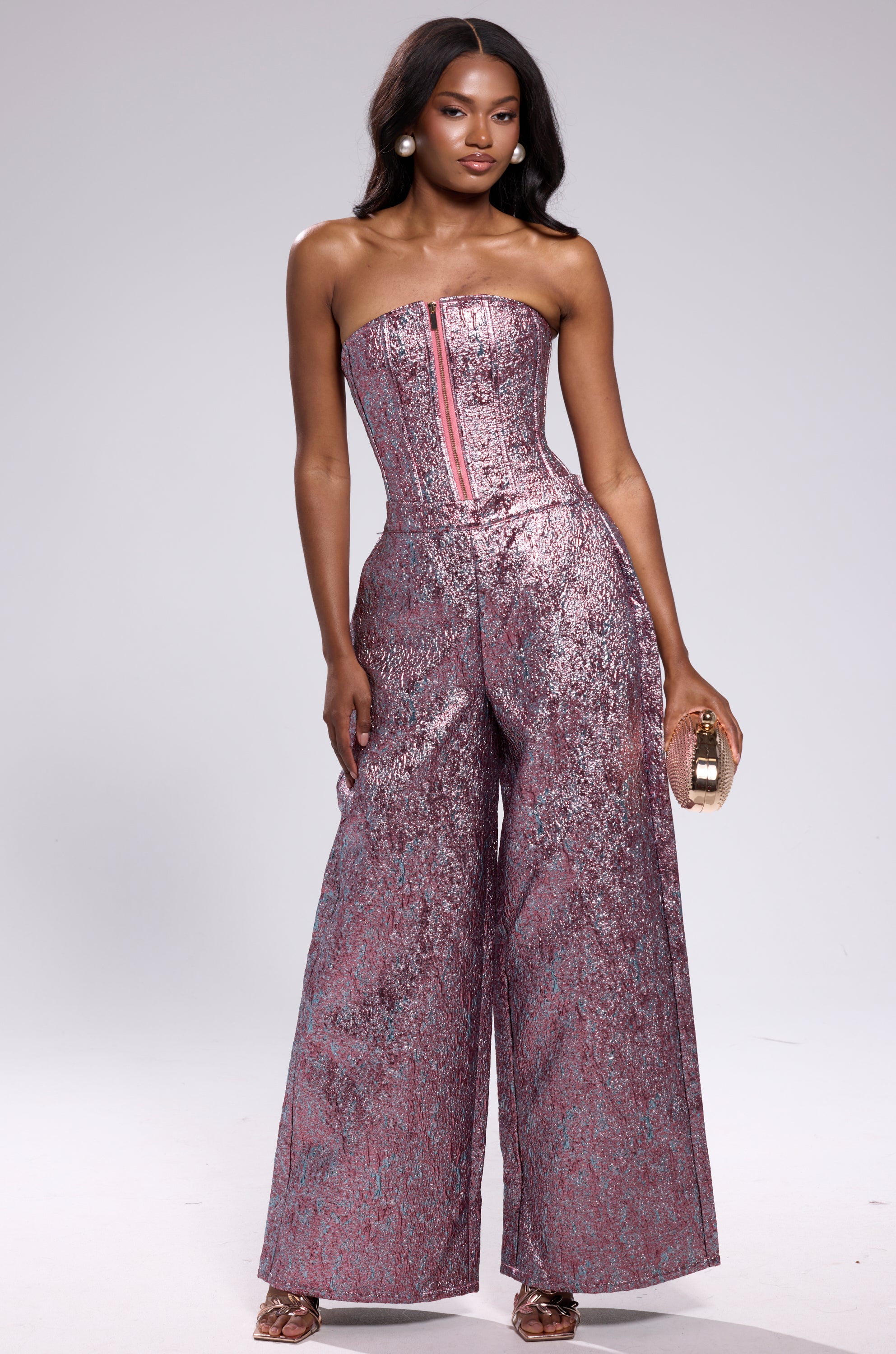 A woman confidently poses in heels against a plain backdrop, wearing NO COMPETITION BROCADE TROUSER—strapless, metallic pink and silver with a textured finish and wide-leg silhouette—paired with a small clutch.
