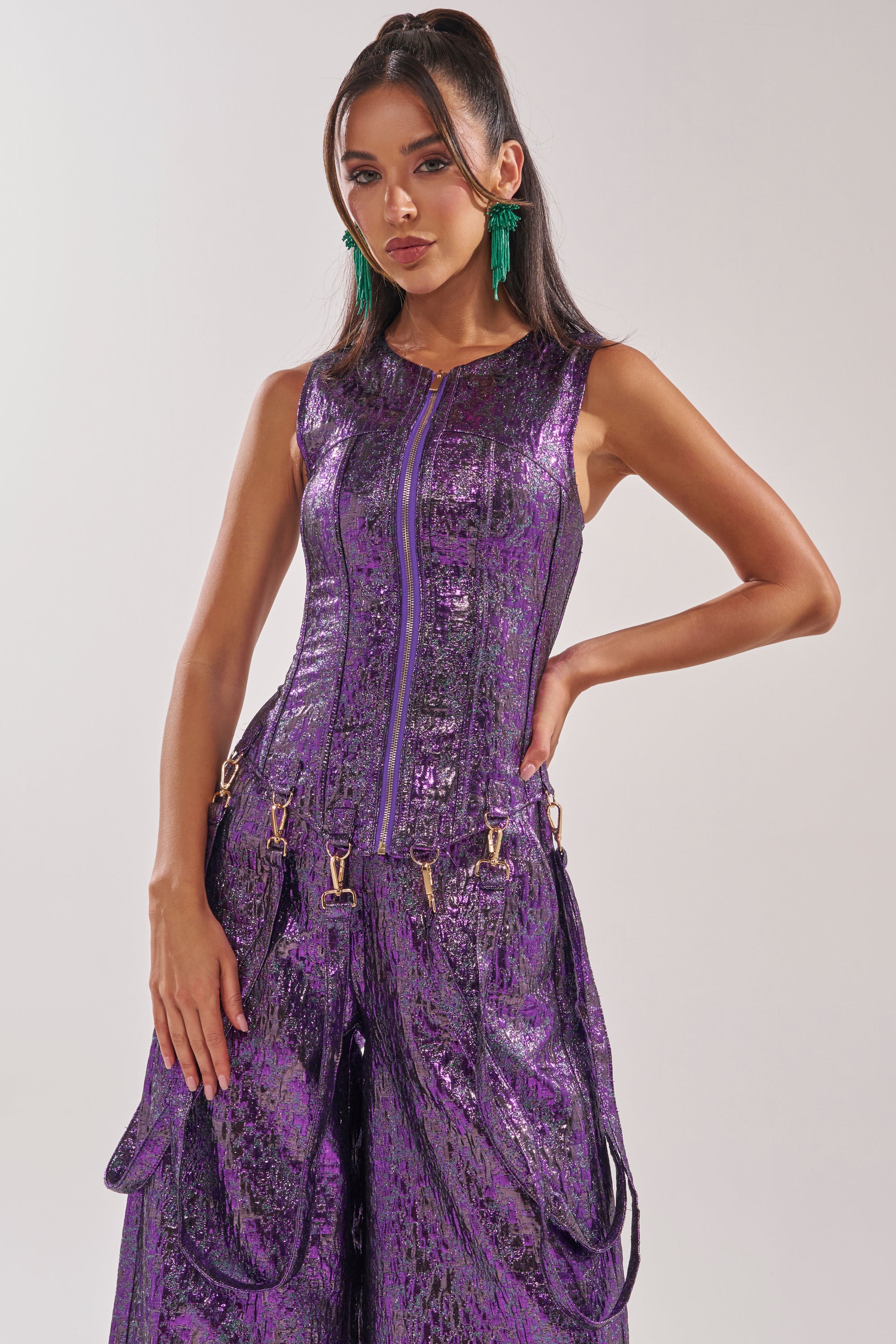 A woman in the ROYALTY BROCADE CORSET TOP, a shiny textured purple piece with zipper and gold hardware, stands before a plain light background. She has long dark hair, green earrings, and a confident look. 0