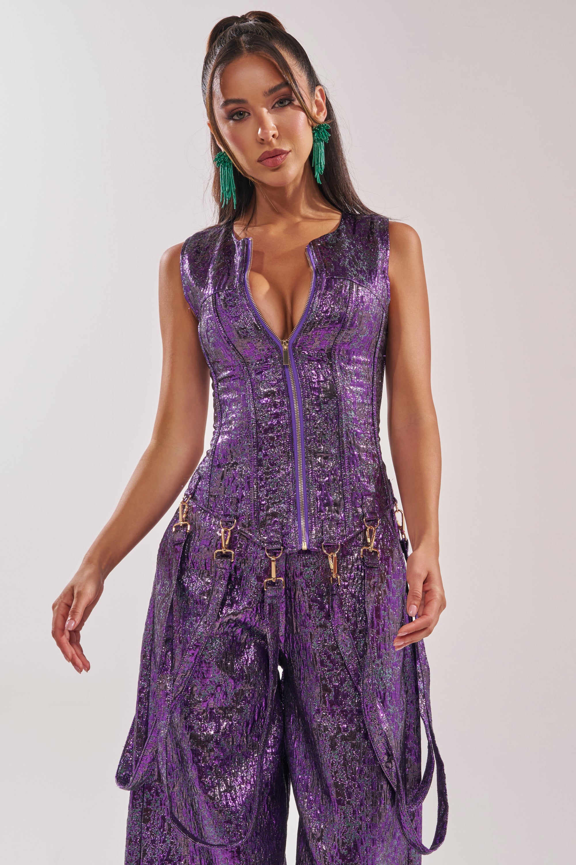 A woman poses against a plain background wearing the ROYALTY BROCADE CORSET TOP in deep purple with gold buckle details, green statement earrings, a zipper front, and a waist-cinching corset silhouette. 1