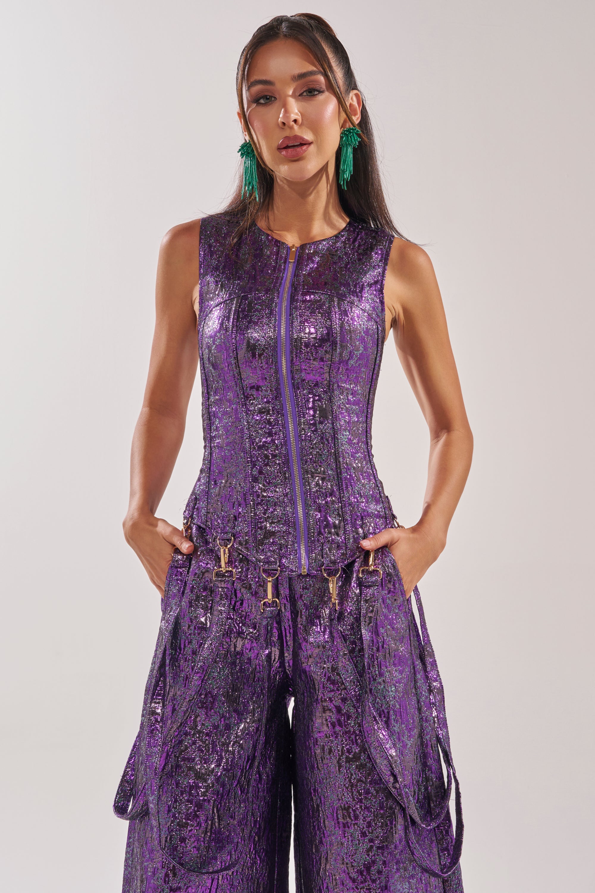 A woman stands against a plain background, wearing the ROYALTY BROCADE CORSET TOP in purple with a shiny texture and front zipper, paired with long green statement earrings and her hands in her pockets. 2