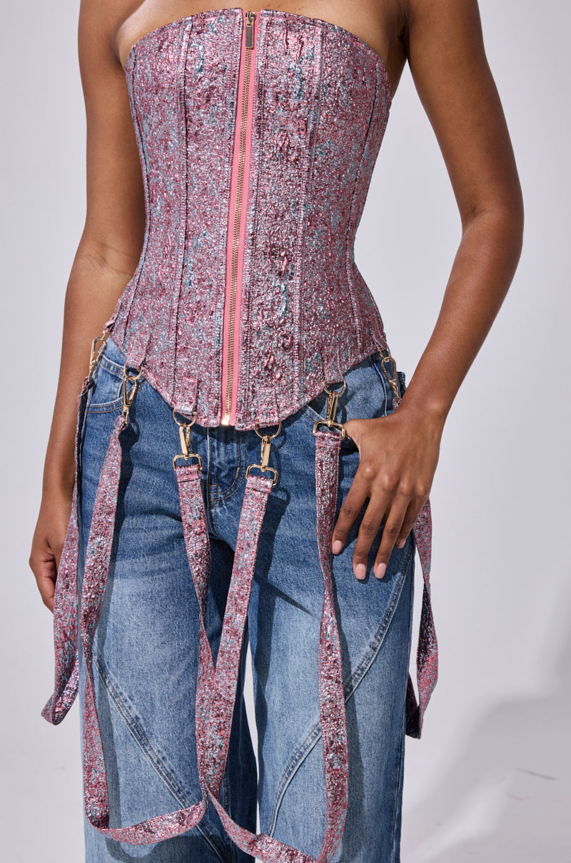 A person wears the NO COMPETITION BROCADE CORSET, a pink textured lace-up top with front zipper and harness straps, styled with loose blue jeans. The image is cropped at the shoulders and mid-thighs.