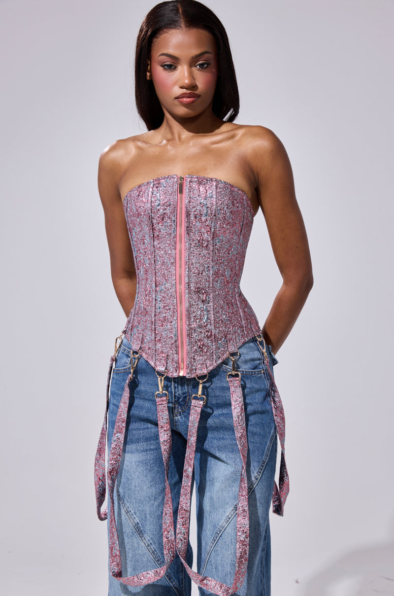 A woman faces forward wearing the NO COMPETITION BROCADE CORSET in pink, featuring a strapless design, front zipper, and attached garter straps, paired with loose blue jeans against a plain light background.