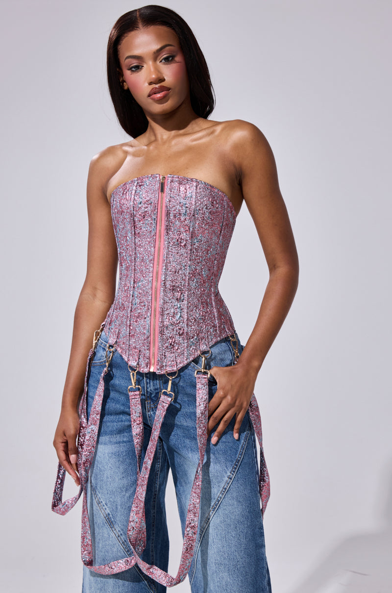 A woman models the NO COMPETITION BROCADE CORSET, featuring metallic brocade fabric with a zipper and suspender-style straps, paired with wide-leg blue jeans against a plain light gray background.