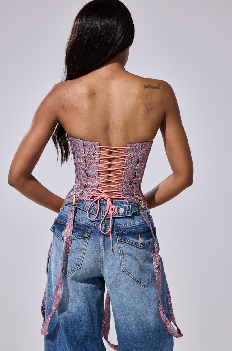 A woman faces away, wearing the NO COMPETITION BROCADE CORSET in pink and purple with ribbon ties, paired with light blue high-waisted jeans. Her long dark hair is down, revealing a small tattoo on her upper back.