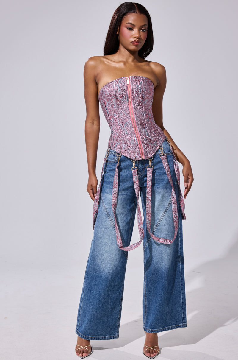 A woman wears the NO COMPETITION BROCADE CORSET, a strapless pink piece with a front zipper, paired with baggy blue jeans and matching pink patterned suspenders. She poses against a plain light background with straight hair.