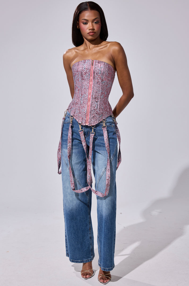 A woman wears the NO COMPETITION BROCADE CORSET, strapless with attached harness straps, paired with loose blue jeans and strappy heeled sandals, standing against a plain light gray background.