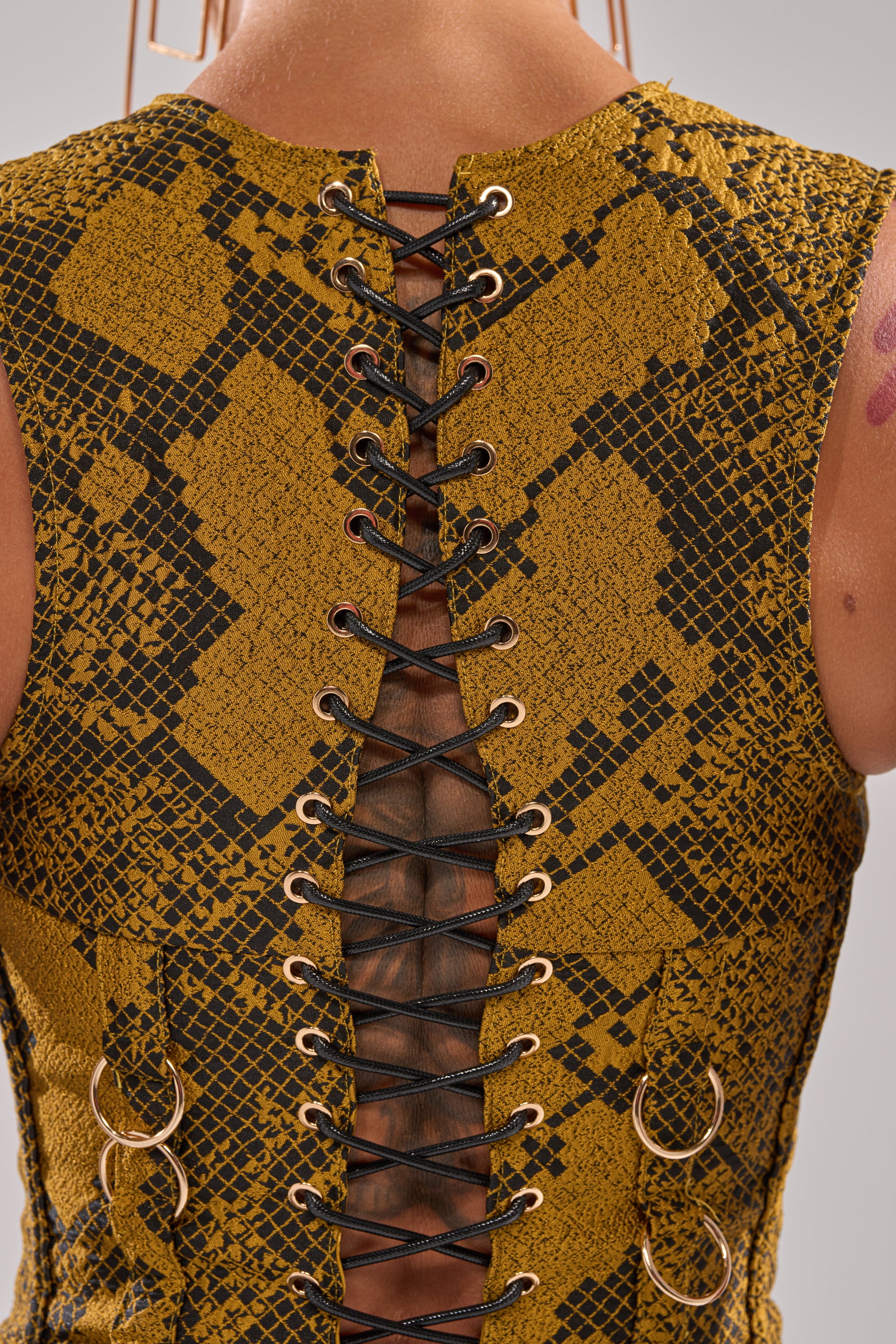 Close-up of a back wearing the SEXY LITTLE THANG SNAKE CORSET, a sleeveless yellow and black jacquard piece with a lace-up panel and metal rings down the center, revealing skin through the striking snake-patterned lacing.
