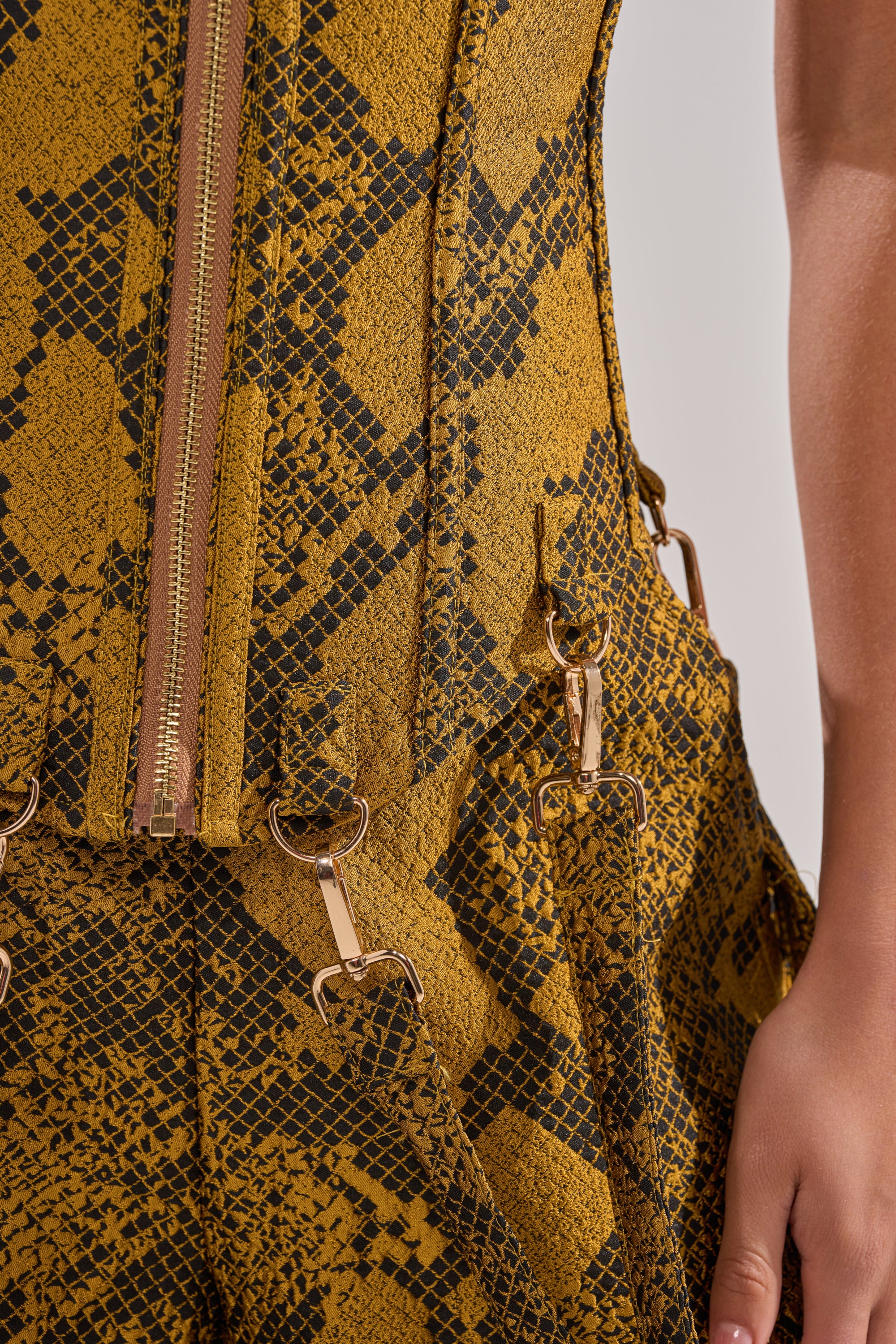 A close-up of the SEXY LITTLE THANG SNAKE CORSET showcases its yellow and black snakeskin-patterned jacquard fabric, zip closure, metal rings, and straps, highlighting the intricate details and a portion of an arm.