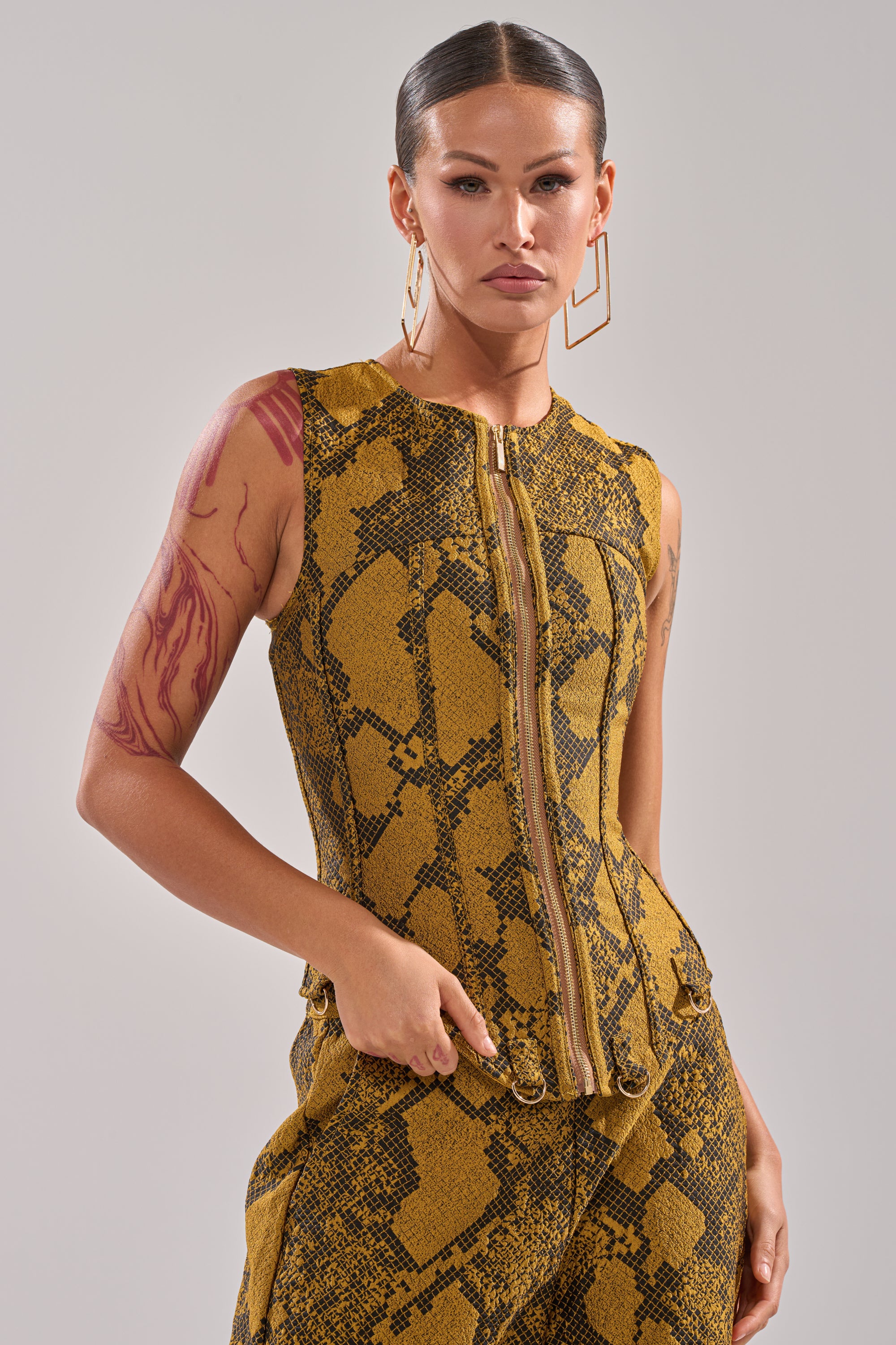 A woman with slicked-back hair and geometric earrings wears the SEXY LITTLE THANG SNAKE CORSET—a yellow and black, snakeskin-patterned sleeveless top—with matching pants, showing off her arm tattoo as she stands confidently against a plain background.