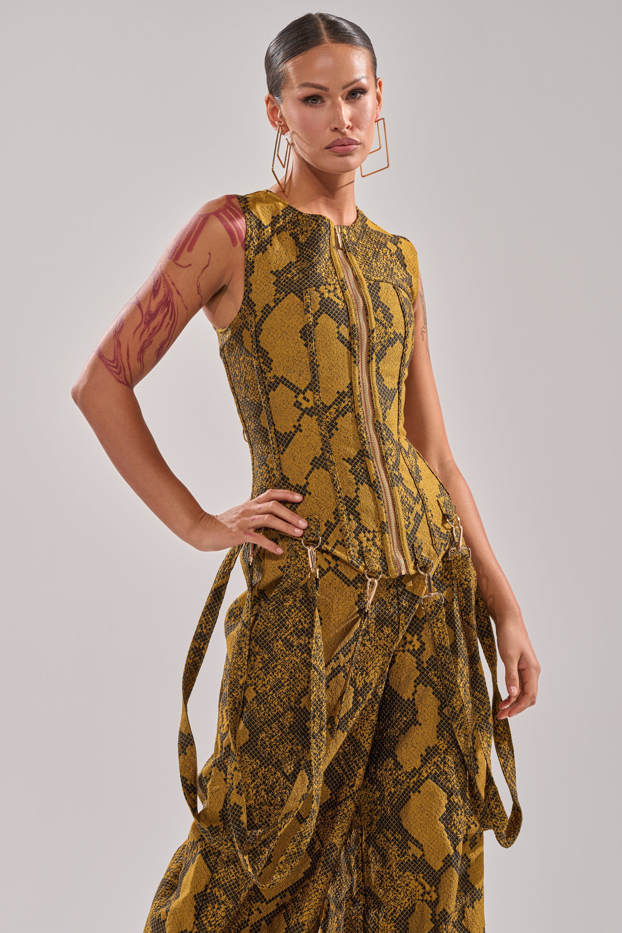 A woman confidently poses with one hand on her hip, wearing the SEXY LITTLE THANG SNAKE CORSET in yellow and brown snakeskin print, paired with wide-legged pants and bold geometric earrings, showing visible arm tattoos against a plain background.