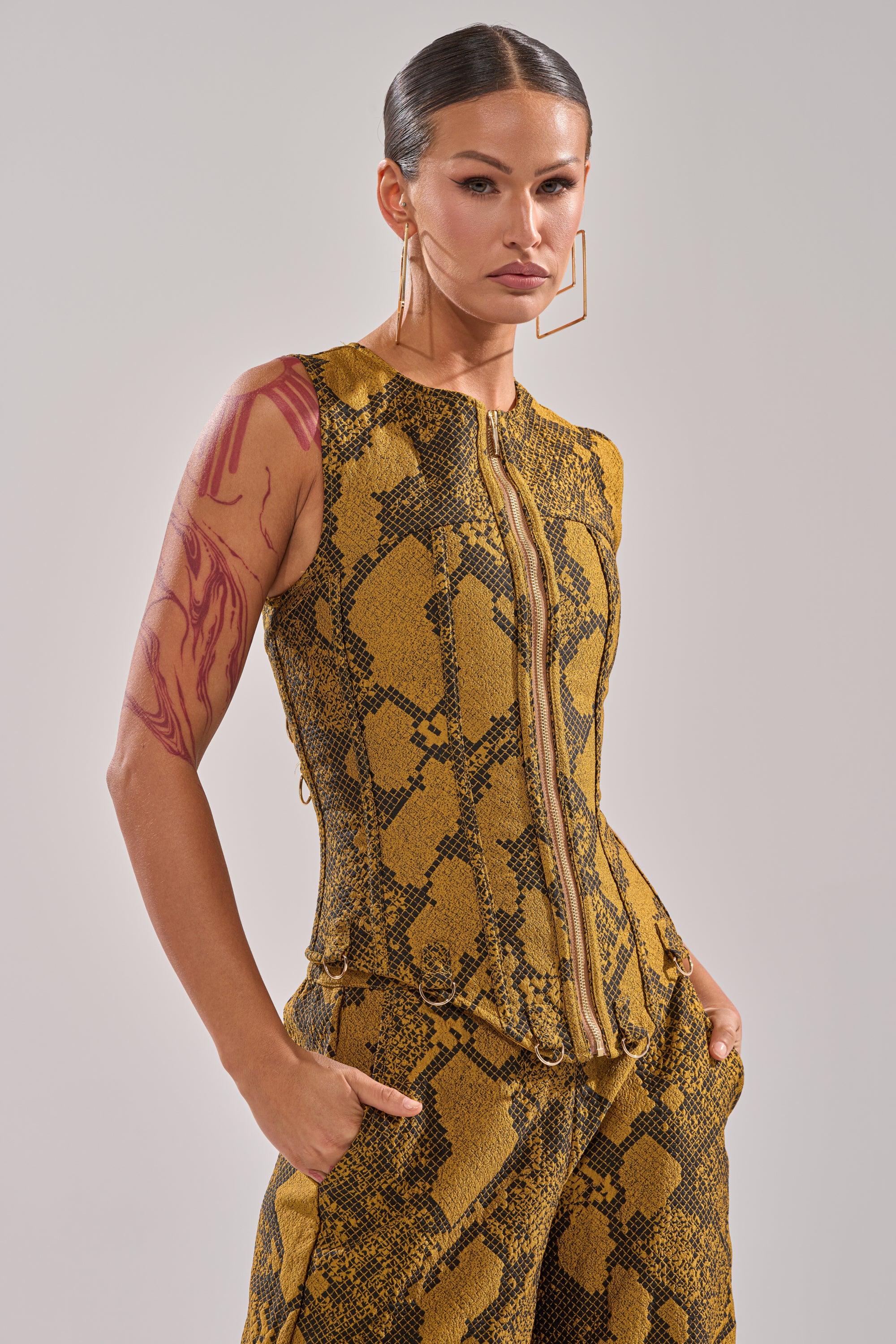 A woman with slicked-back hair poses confidently in the SEXY LITTLE THANG SNAKE CORSET and matching pants, accessorized with geometric gold earrings and a visible red tattoo for a bold snakeskin-patterned look.