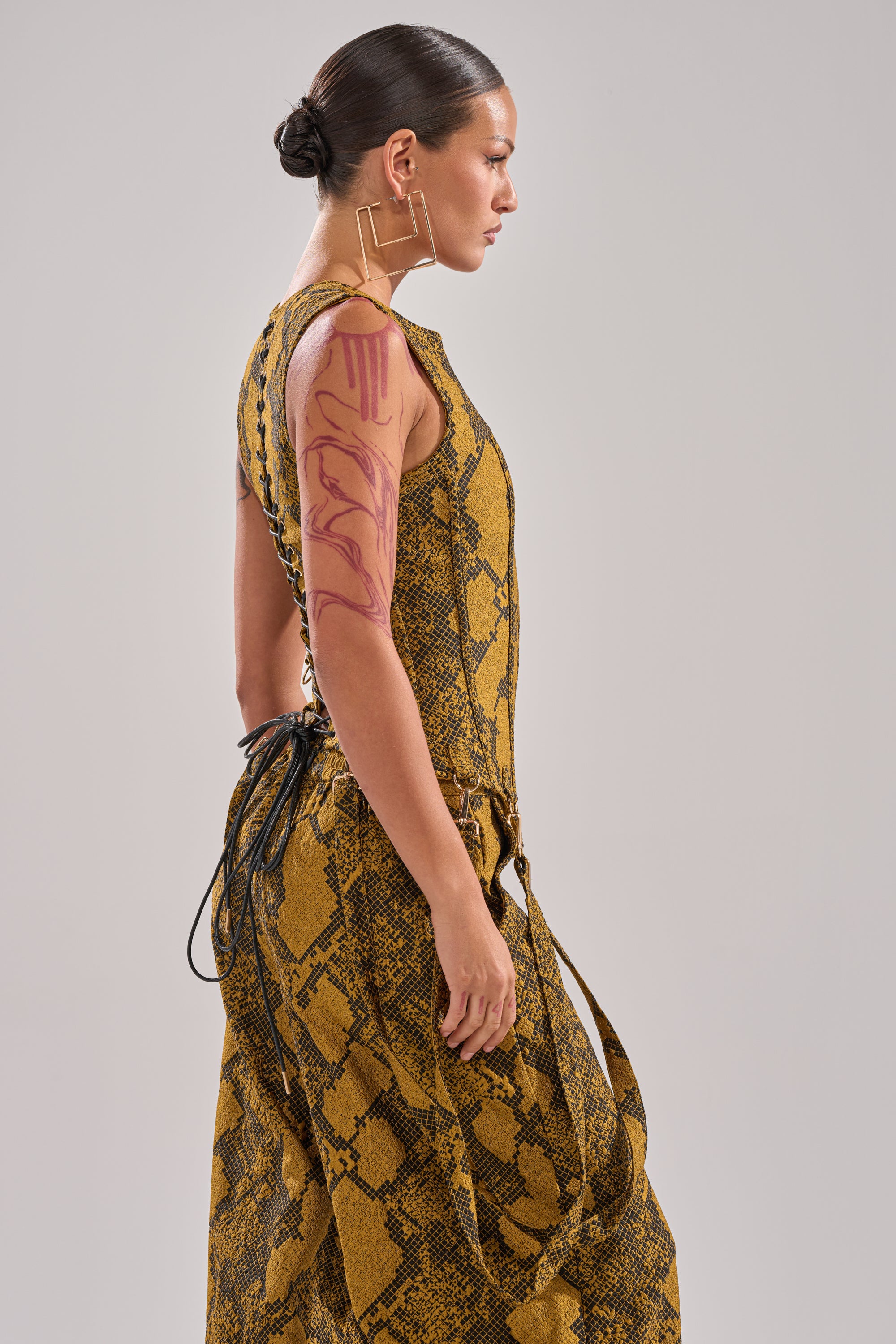 A woman with dark hair in a bun wears a sleeveless, yellow SEXY LITTLE THANG SNAKE CORSET dress. She stands sideways against a plain background, accessorized with geometric gold earrings and a red tattoo on her upper arm.