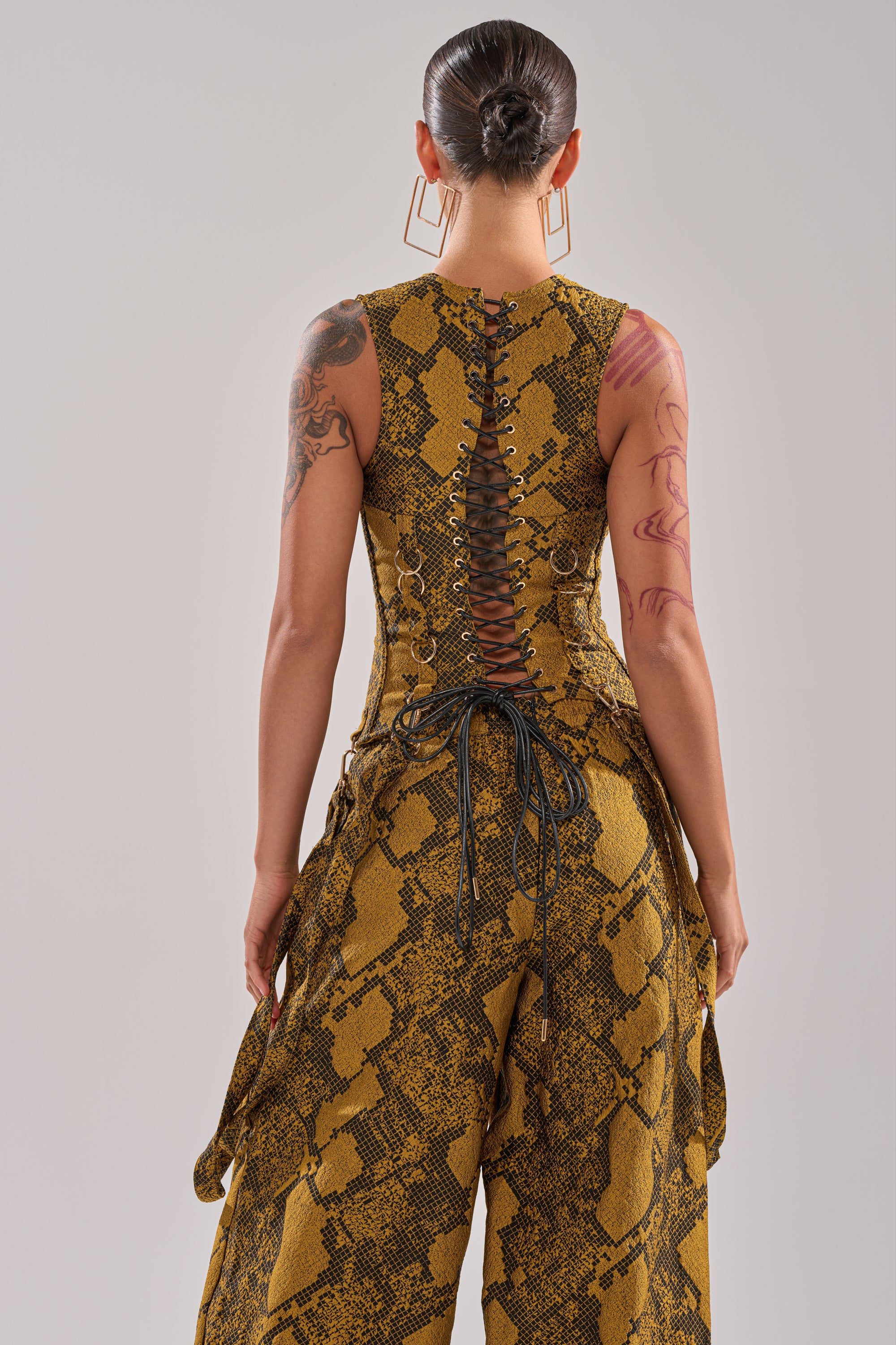 A person with a sleek bun and large hoops wears the SEXY LITTLE THANG SNAKE CORSET—a yellow and brown snakeskin-patterned sleeveless piece with a lace-up back and sash details. Tattoos are visible on their arms.
