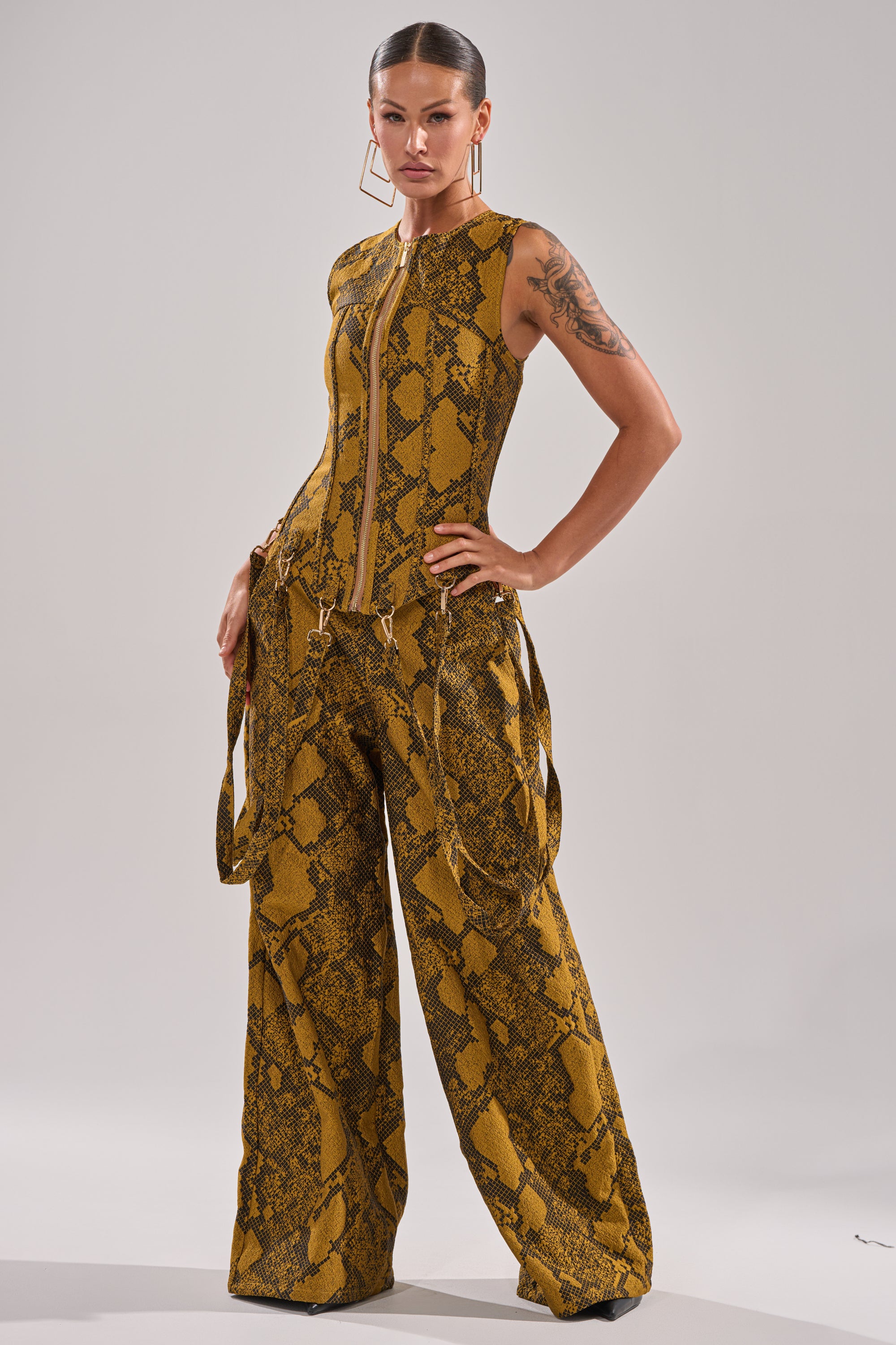 A woman poses confidently in the SEXY LITTLE THANG SNAKE TROUSER—a sleeveless, zip-up jumpsuit with wide-leg trousers and a yellow and brown snakeskin print—styled with slicked-back hair, hoop earrings, and an arm tattoo.