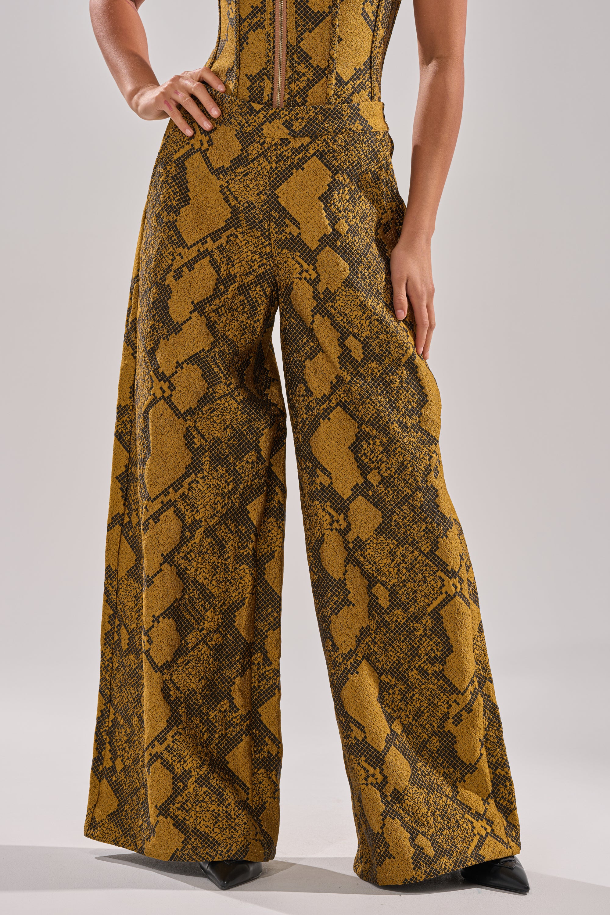 A person models the SEXY LITTLE THANG SNAKE TROUSER, featuring striking yellow and brown high-rise, wide-leg snakeskin pants. Only their lower torso and legs are shown, with hands on hips.