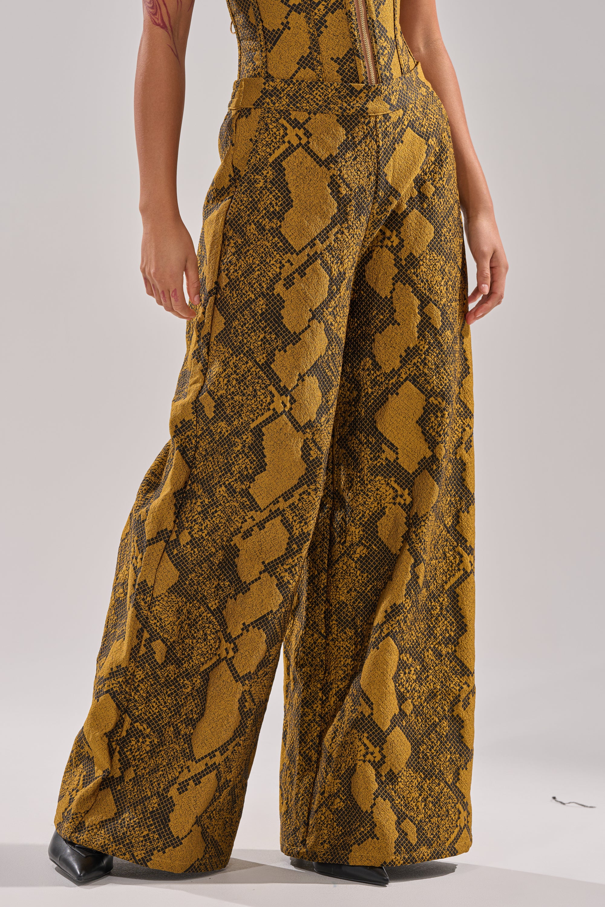 A person wears the SEXY LITTLE THANG SNAKE TROUSER, featuring a high waist and wide legs with a yellow and black snakeskin print, standing with one hand by their side. The image is cropped from the shoulders down.