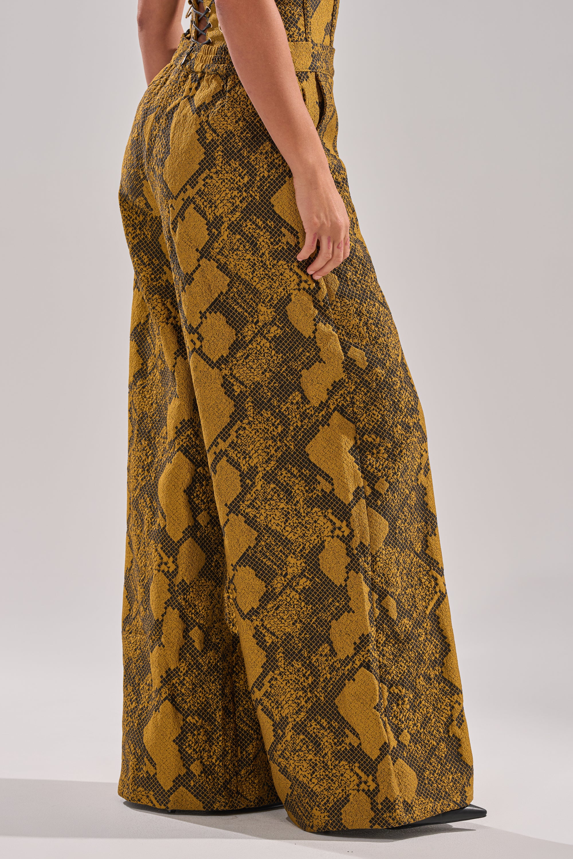 A person models the SEXY LITTLE THANG SNAKE TROUSER, featuring high-rise, wide-leg design with a yellow and brown snakeskin pattern, standing against a plain light background. Only the lower body is shown.