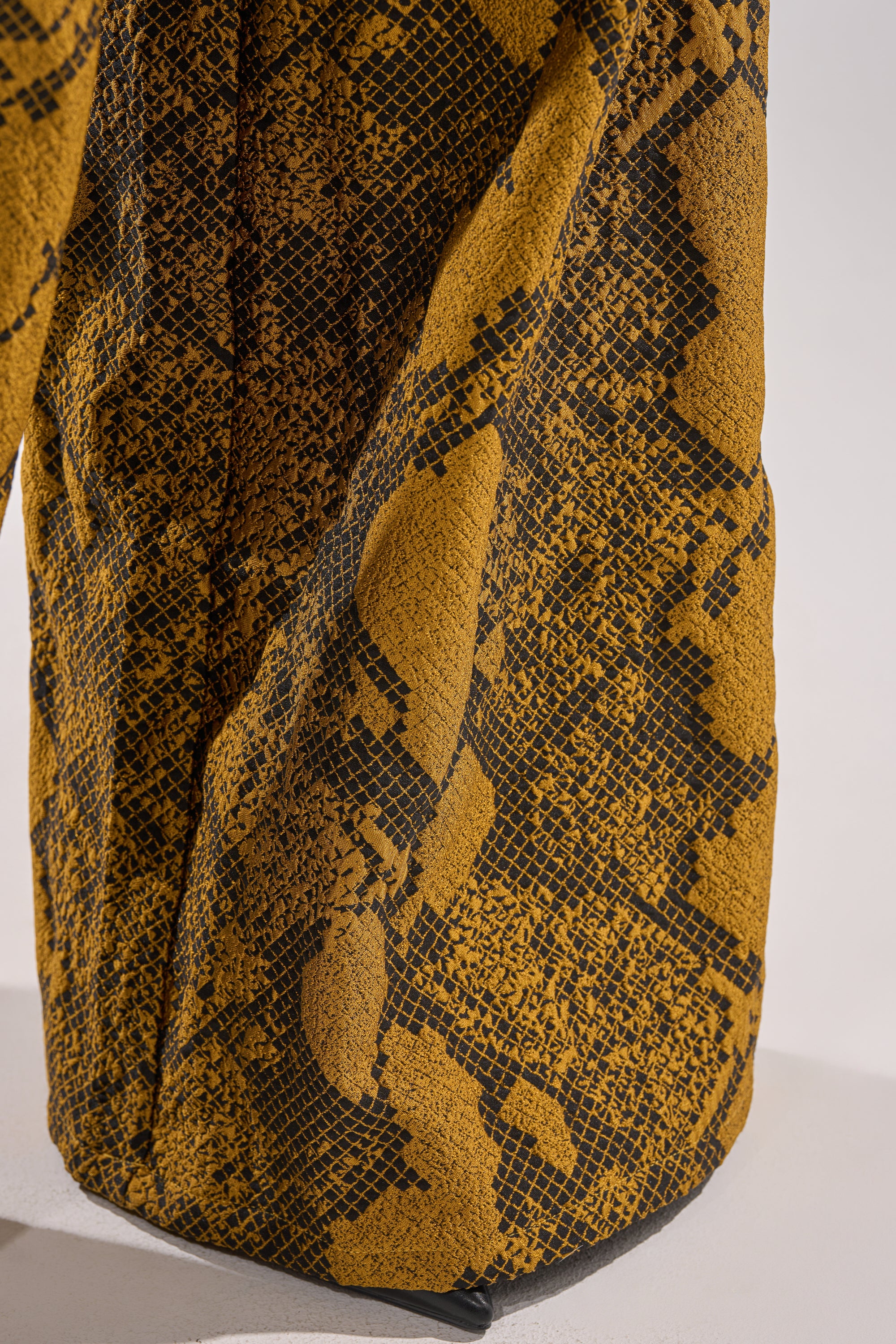 Close-up of yellow fabric with a black, snake-skin-like grid pattern, similar to the SEXY LITTLE THANG SNAKE TROUSER. The draped material forms textured folds and shadows against a plain background.